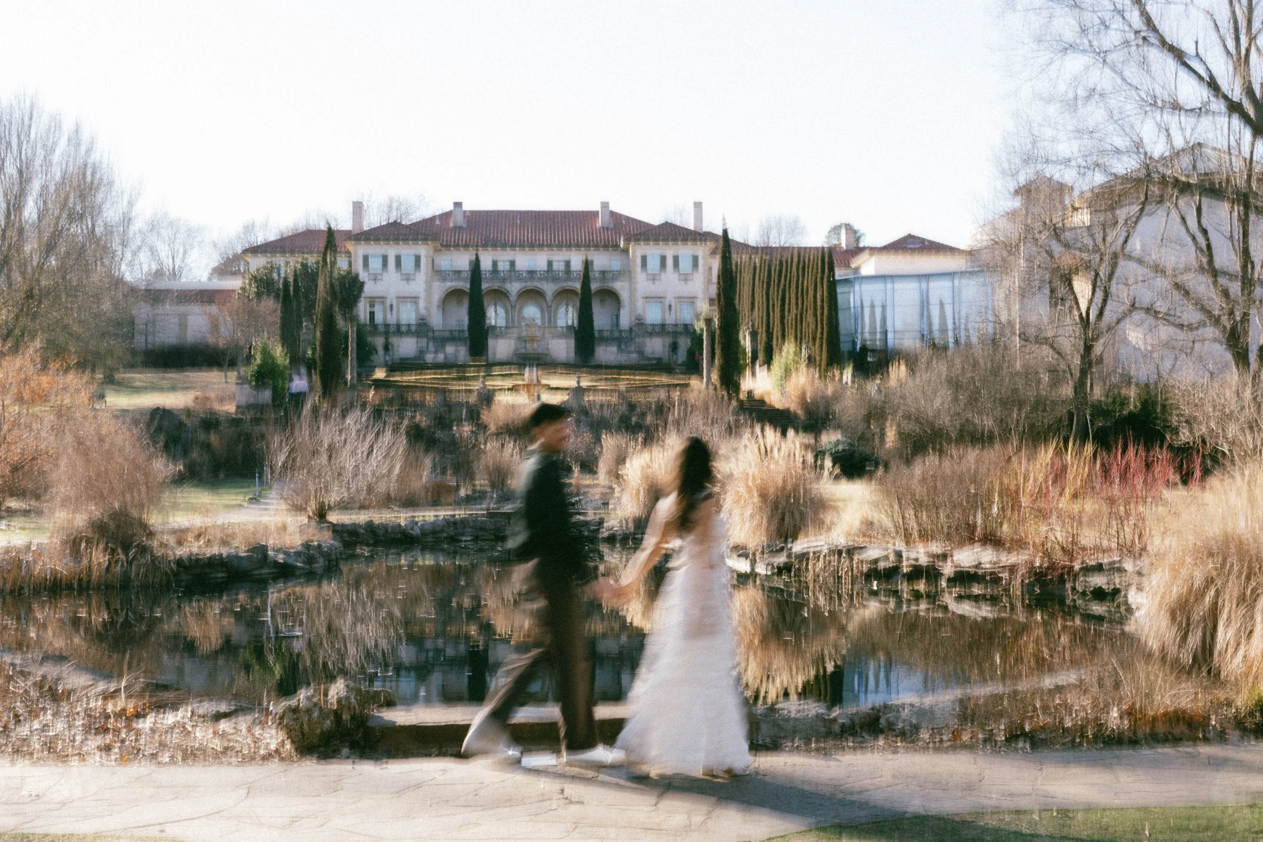 Cinematic wide shot at Philbrook Museum Tulsa. Couple blurred in foreground with clear view of the historic mansion and expansive gardens, capturing a relaxed luxury wedding vibe on film