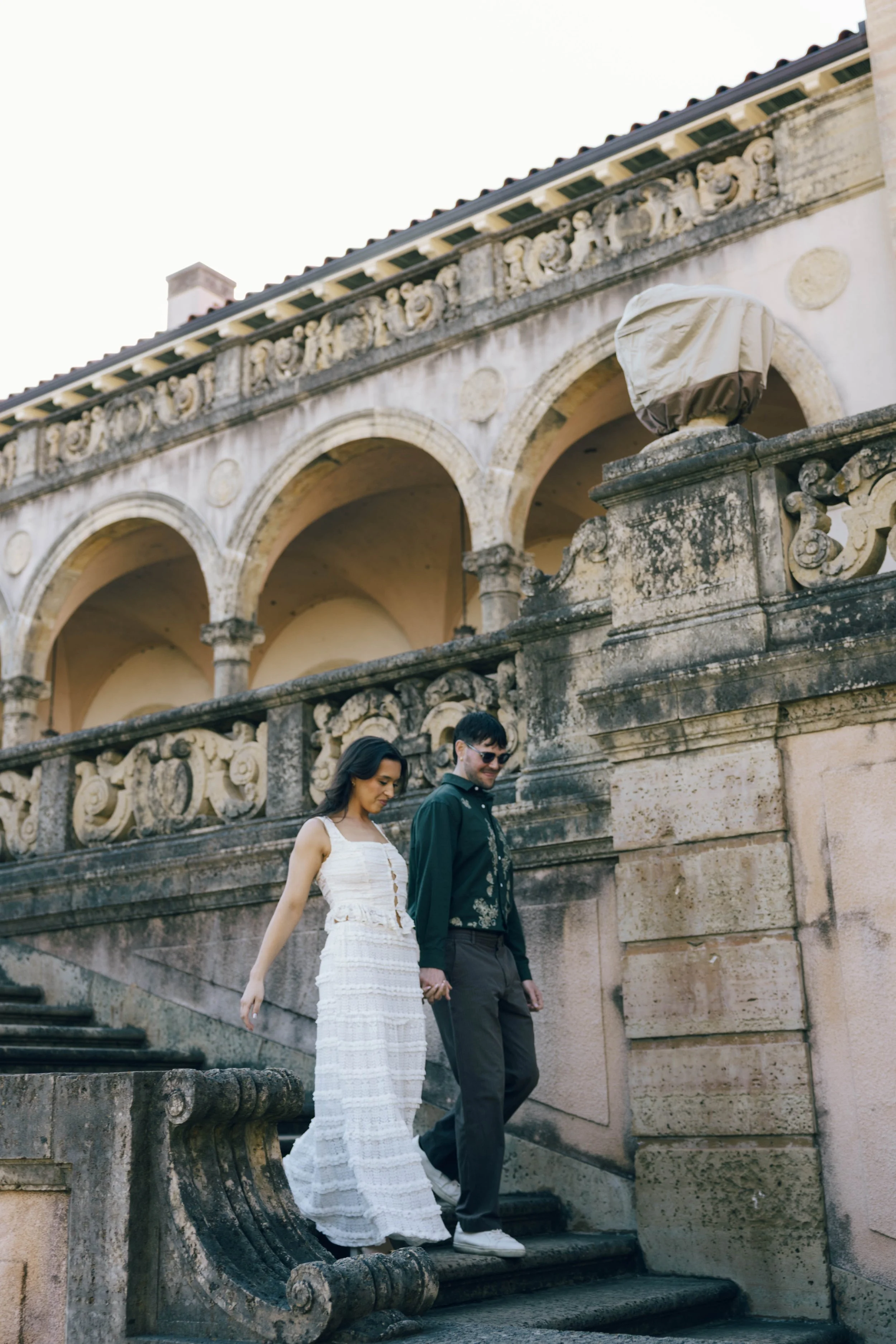 Engagement session photography at Philbrook Museum Tulsa featuring the iconic stone loggia and outdoor staircase. The couple walks through a scene defined by grand arches and old-world textures, captured with a cinematic, film-inspired aesthetic