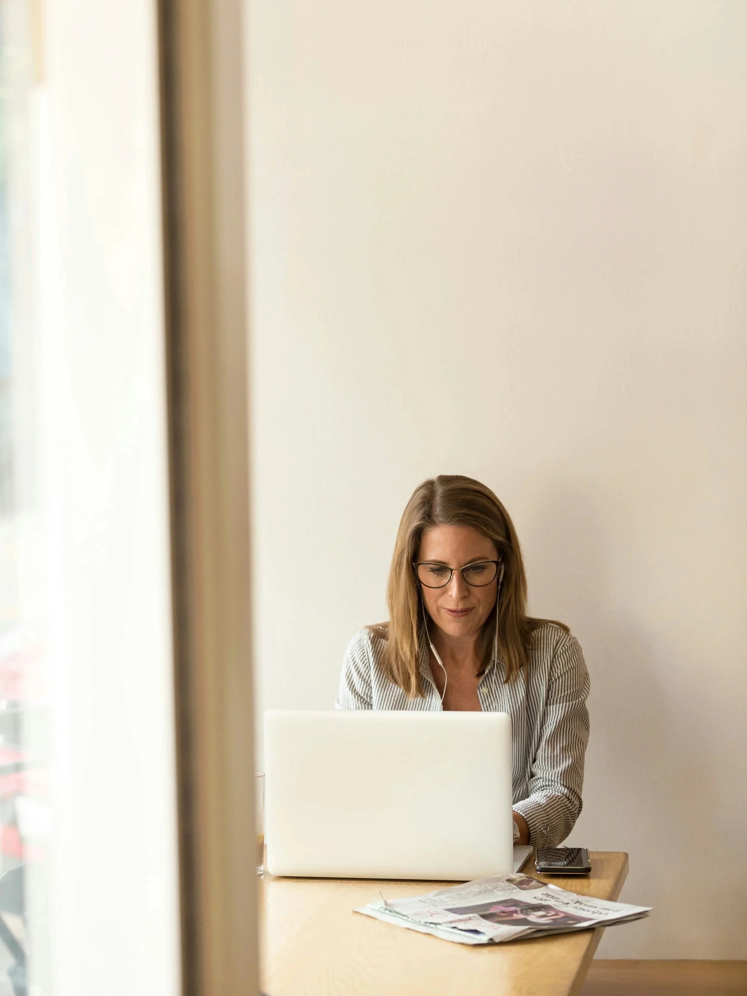 Mujer trabajando en una oficina frente a una laptop sobre una mesa, con diarios y teléfono móvil, y usando gafas.