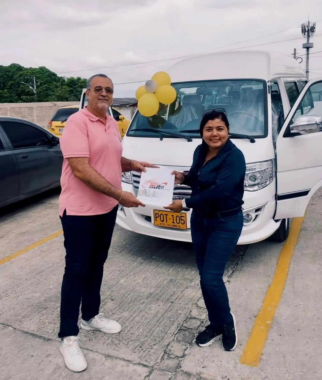 Dos personas, un hombre y una mujer, en una ceremonia de entrega de un vehículo blanco en un estacionamiento. El hombre viste una camisa rosa y la mujer una chaqueta azul, ambos sosteniendo un documento. En el fondo hay varios autos y un cielo nublad