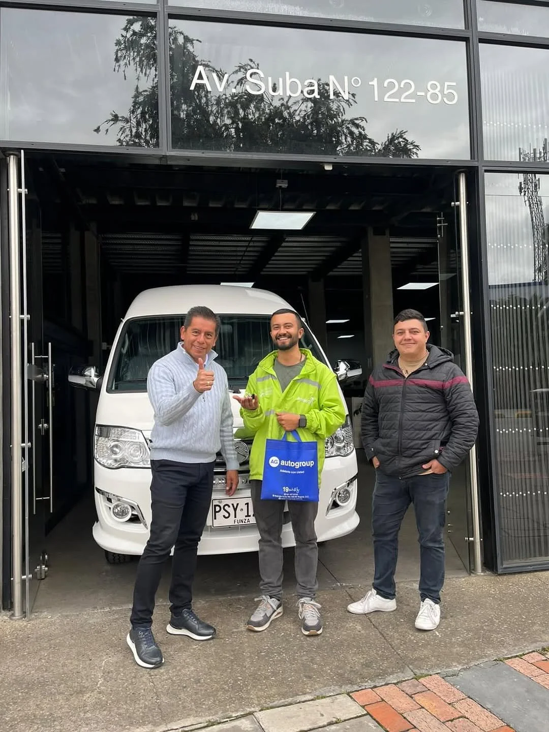 Tres hombres felices posando frente a una camioneta blanca en una tienda de autos. Uno de ellos lleva una bolsa azul y todos están sonriendo.