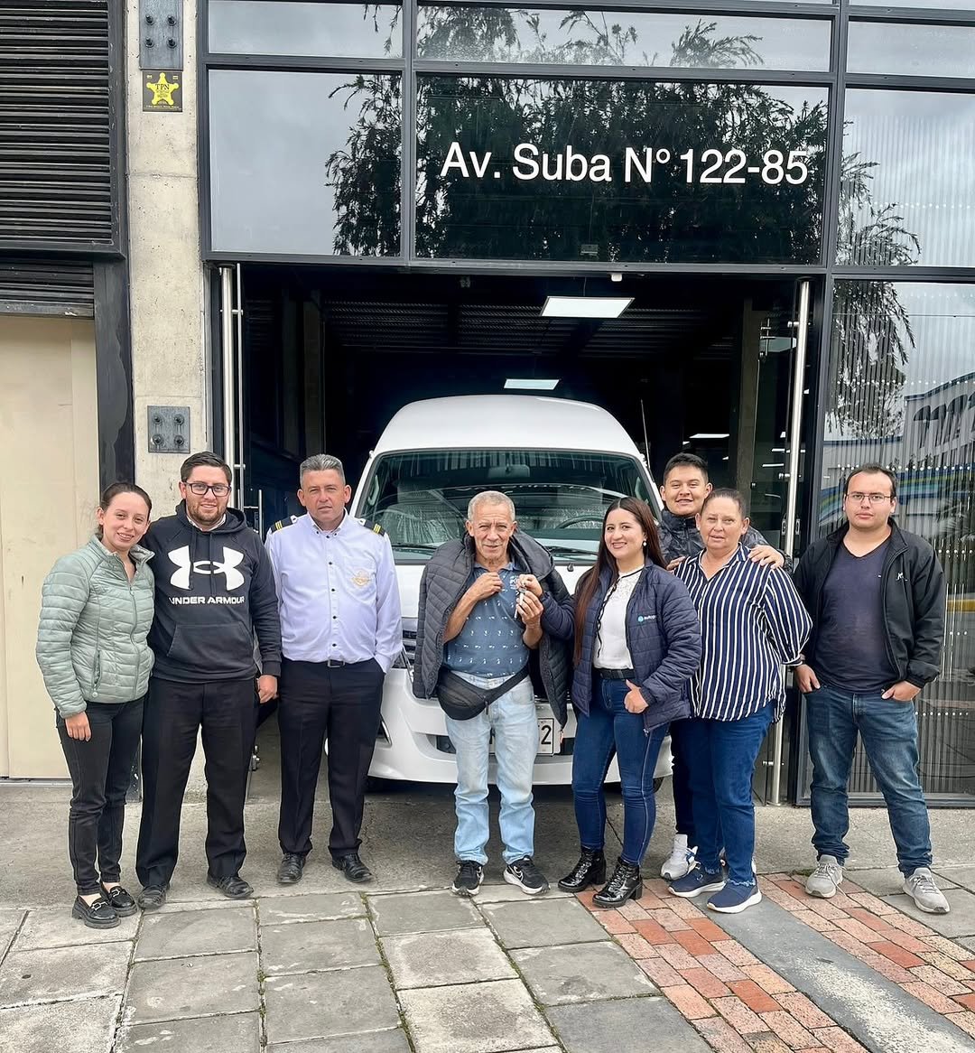 Grupo de personas posando frente a una oficina con un vehículo en el garaje, algunos son empleados y uno parece ser un cliente, en un día nublado.