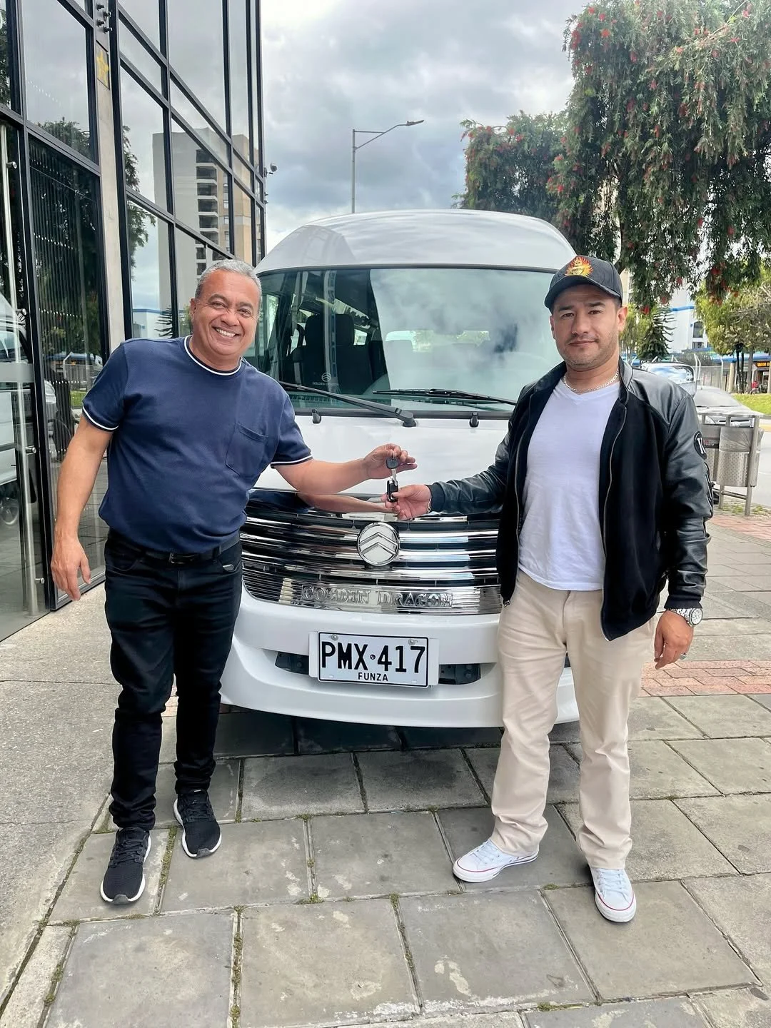 Dos hombres posando frente a un vehículo blanco, uno de ellos entregando unas llaves. Ambos están sonriendo, uno vestido con camiseta azul y pantalones negros, y el otro con sudadera negra y pantalones claros. La imagen fue tomada en un entorno urban