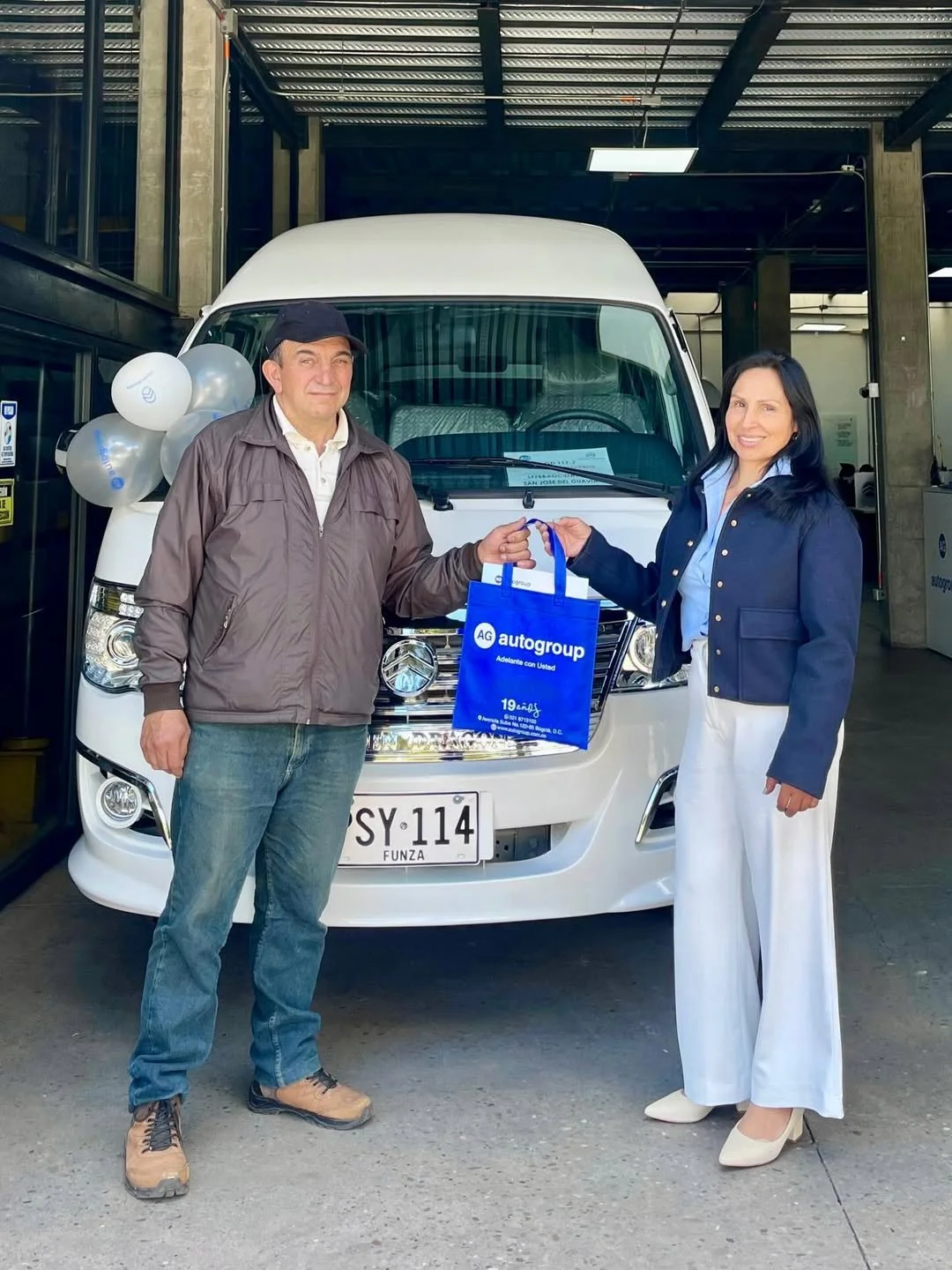 Una mujer y un hombre están en un estacionamiento, con un furgón blanco detrás, la mujer le entrega una bolsa azul a el hombre. Ambos sonríen. La mujer viste un blazer azul y pantalones blancos, el hombre lleva chaqueta gris y gorra. La bolsa azul ti