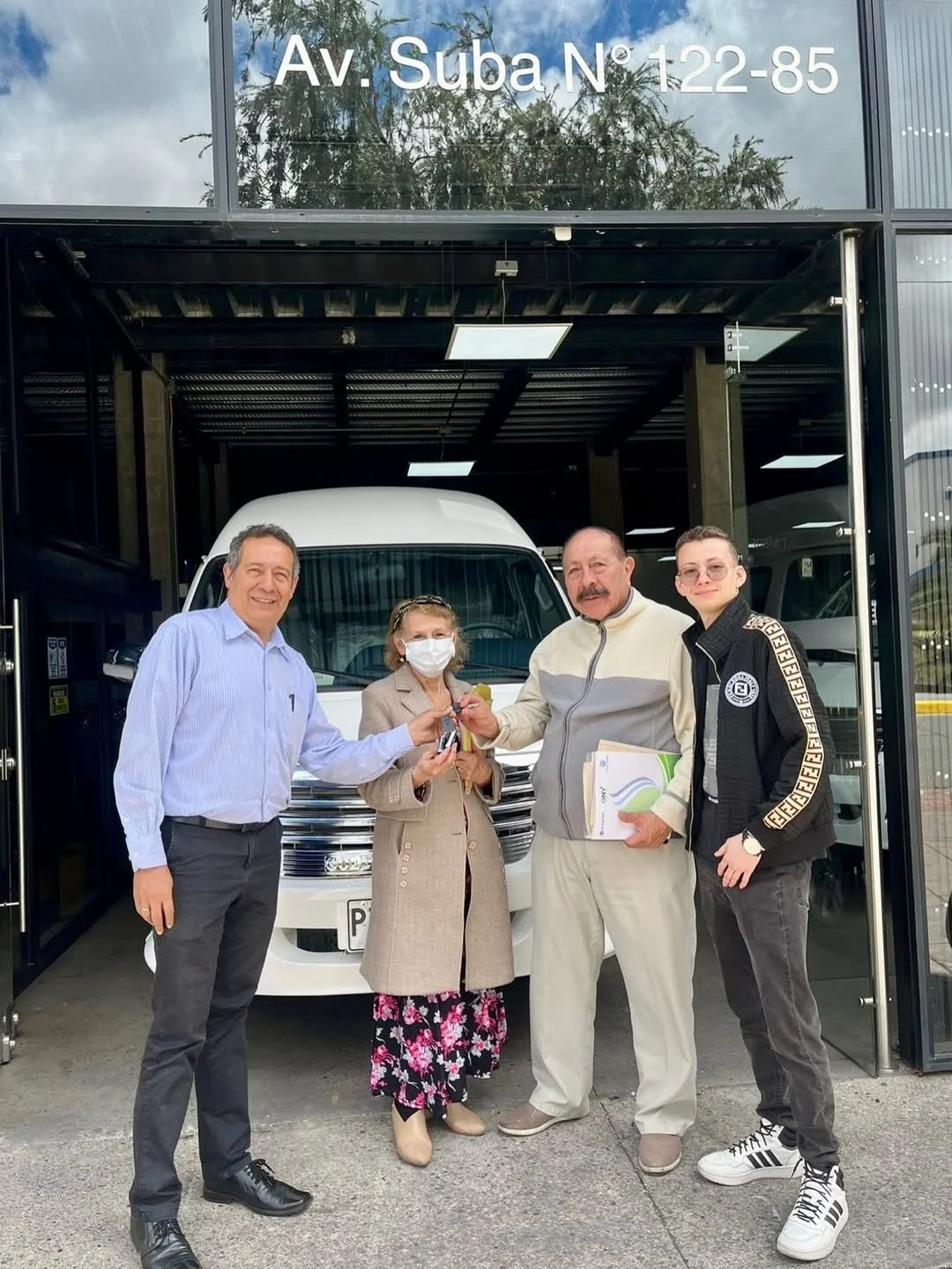 Cuatro personas en la entrada de un concesionario de autos, entregándose las llaves de un vehículo blanco.