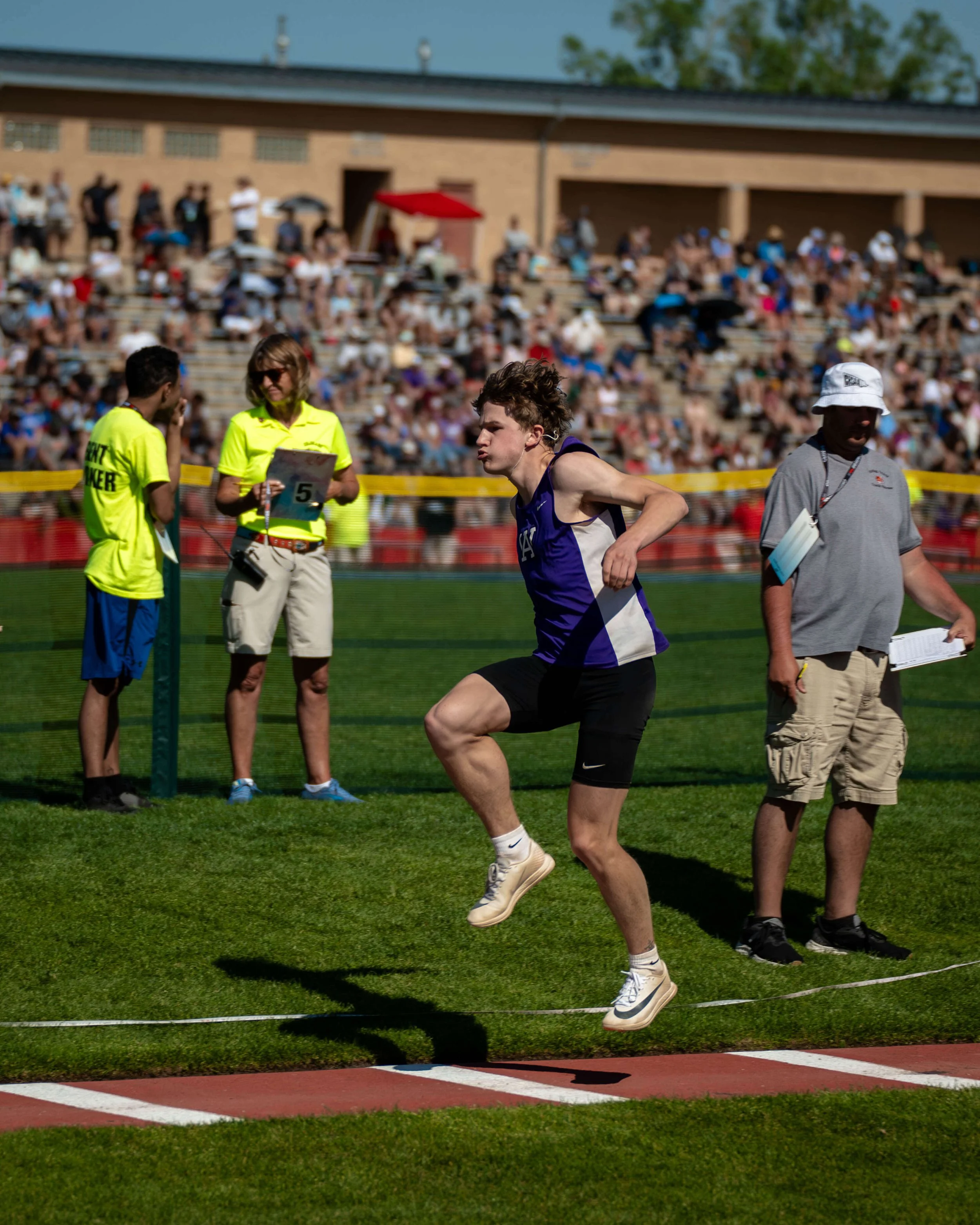 A male track athlete in a purple and black uniform jumping over a hurdle during a race at a stadium with spectators