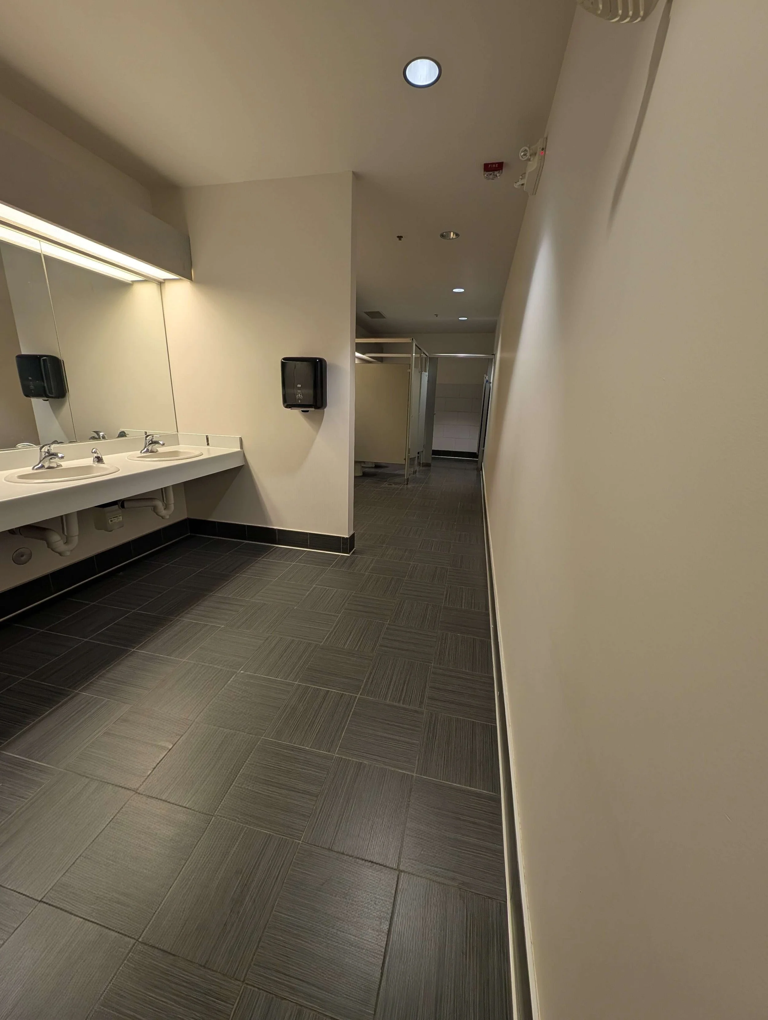 A public restroom with two sinks, a mirror, black soap dispensers, and a hallway leading to bathroom stalls.