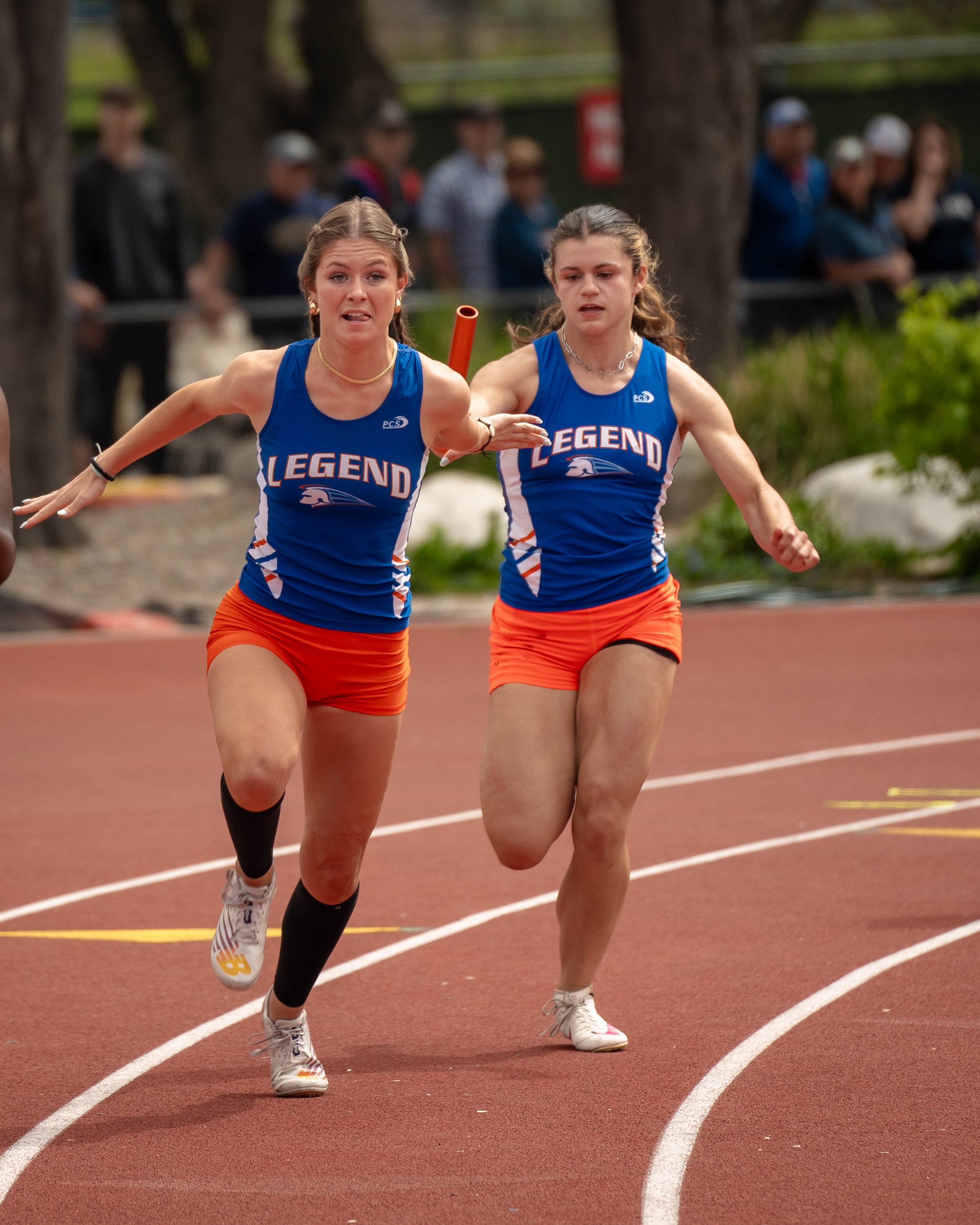 Two female athletes competing in a race, wearing blue and orange uniforms, on a running track with spectators in the background.