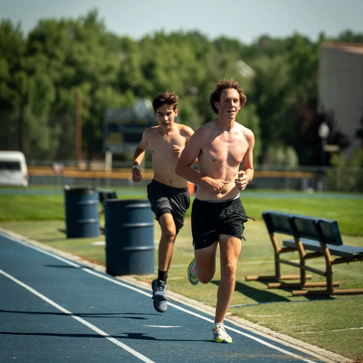 Two shirtless distance men running on a track outdoors during daytime, with trees and benches in the background.