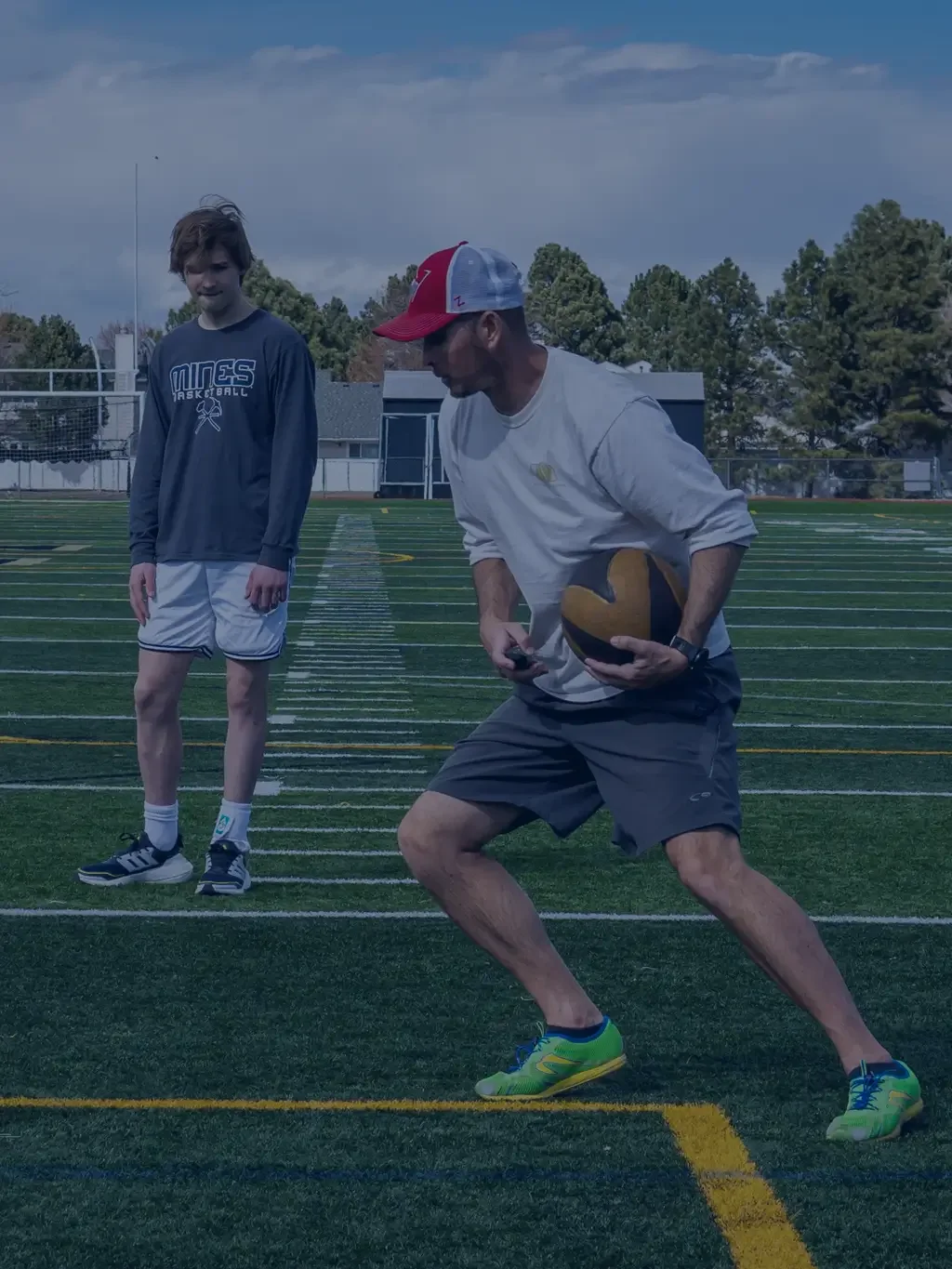 A man in gray shorts, a white shirt, and a red and gray cap is holding a football in a formation on a football field, with a young man in a black long sleeve shirt and white shorts standing nearby, with trees and a building in the background.