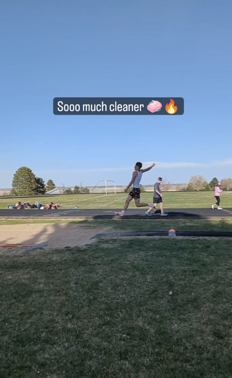 A young man mid flight during a long jump takeoff with a group of people running in the background outdoors under a clear blue sky.