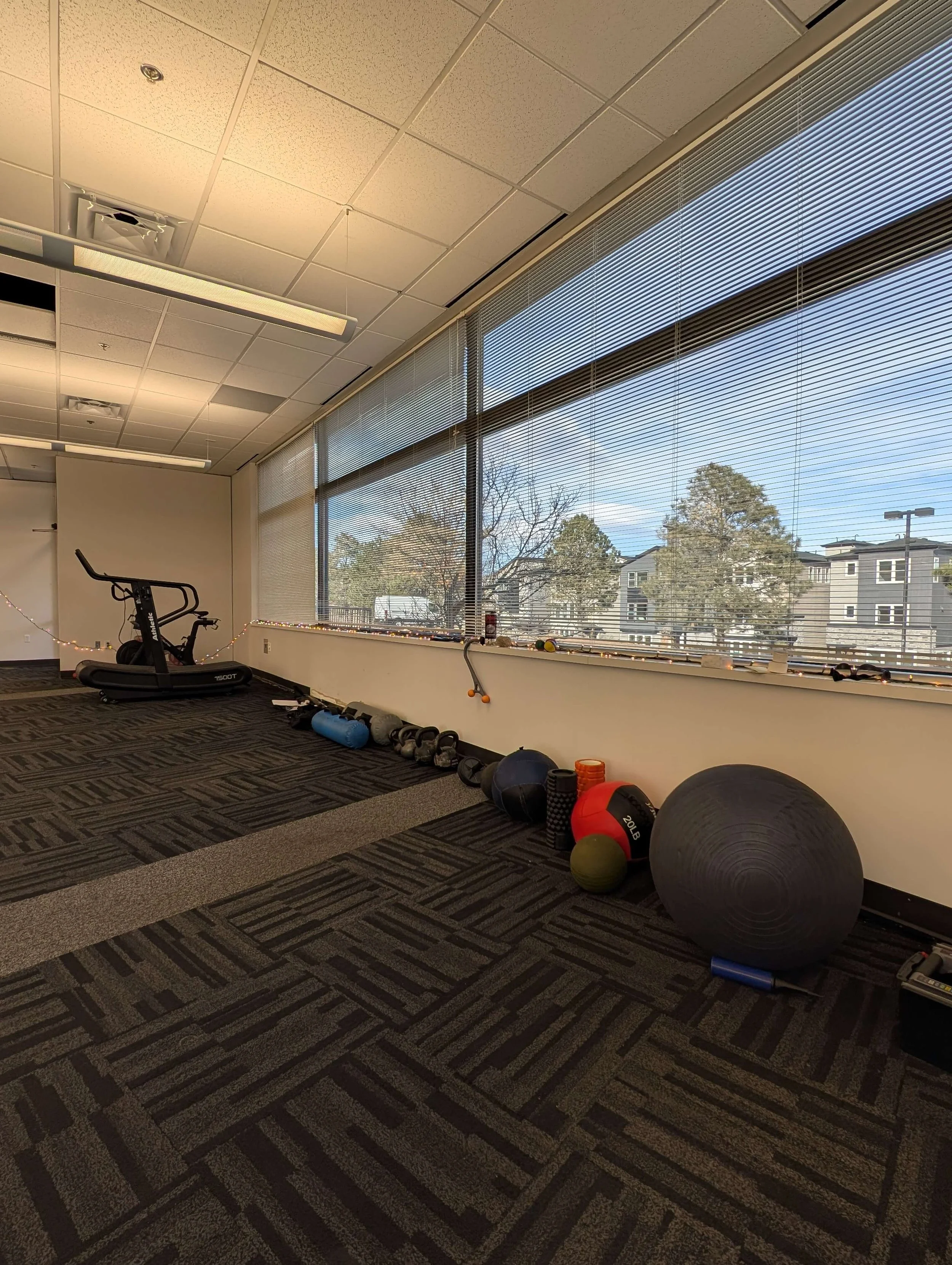 Empty workout room with exercise equipment including a treadmill, stationary bike, kettlebells, medicine balls, foam rollers, and a large exercise ball near windows with blinds, looking out onto a parking lot and trees.