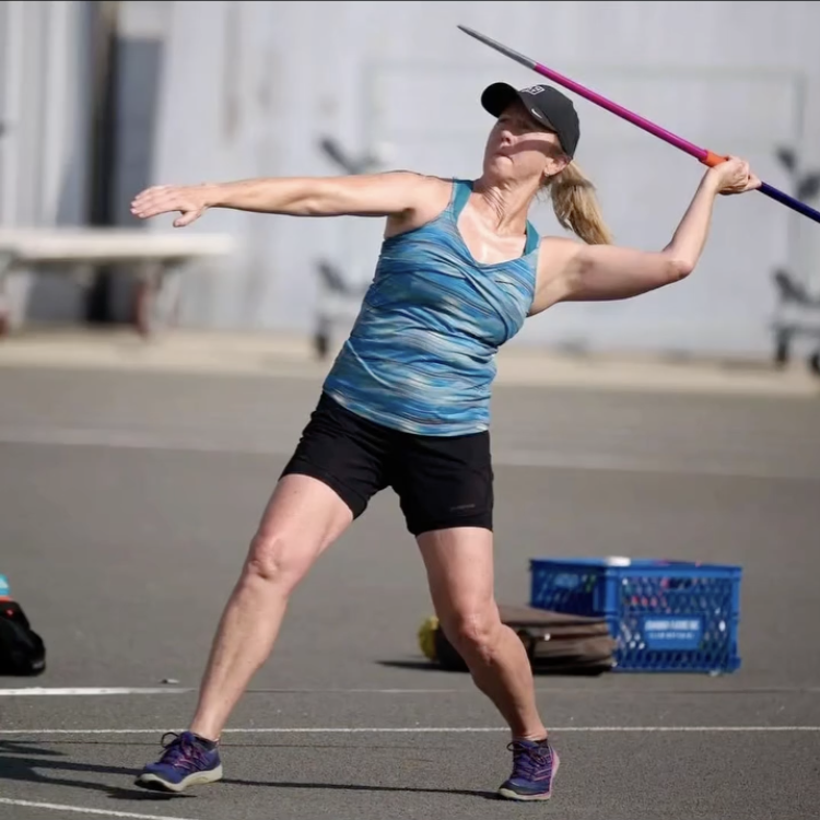 An older woman practicing javelin throw outdoors, with her arm extended back holding a pink and purple javelin, wearing a black cap, blue patterned tank top, black shorts, and purple athletic shoes.