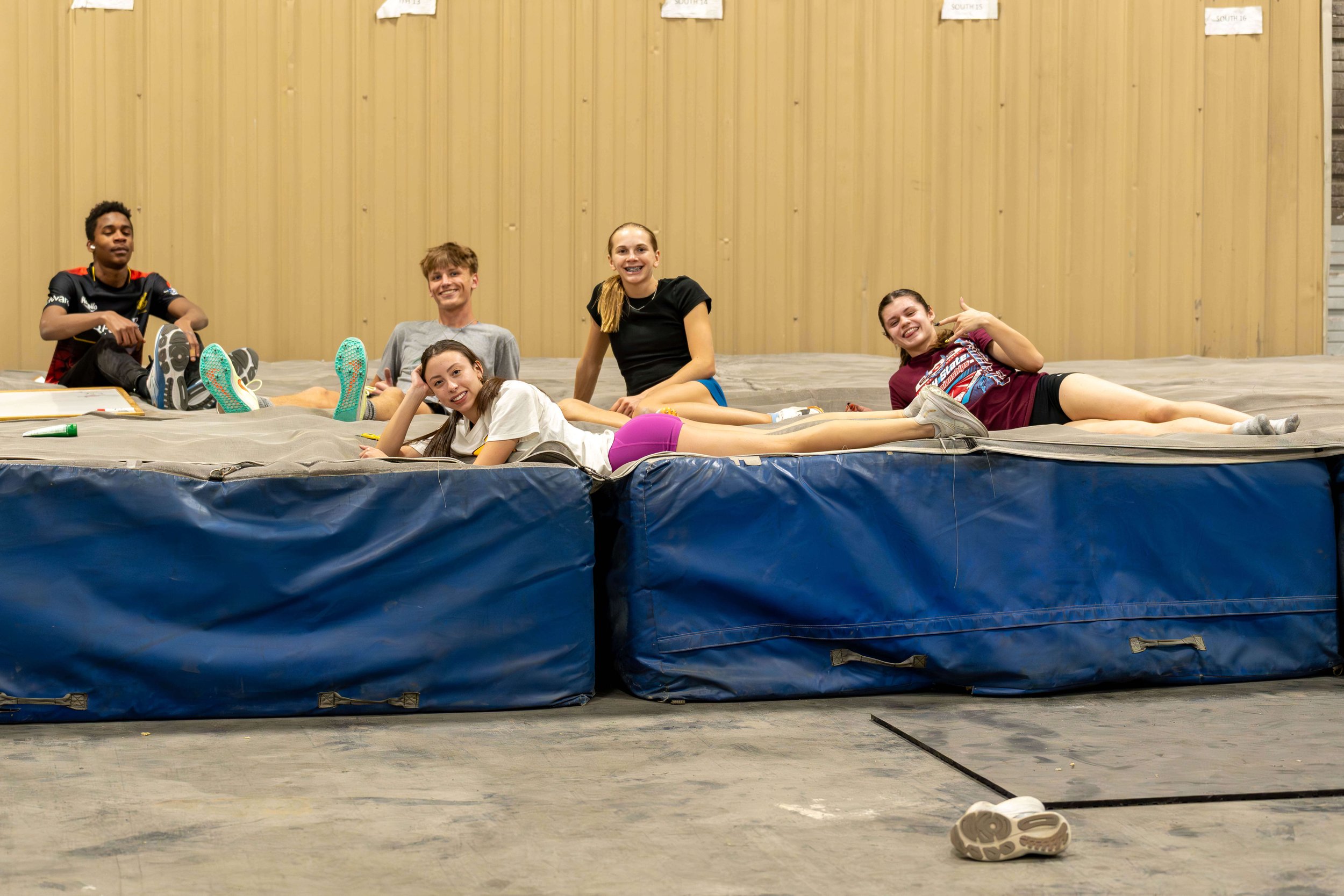 Five young people relaxing on a padded area indoors, with a wooden wall behind them. They are smiling and wearing casual sportswear, with some lying down and some sitting up.