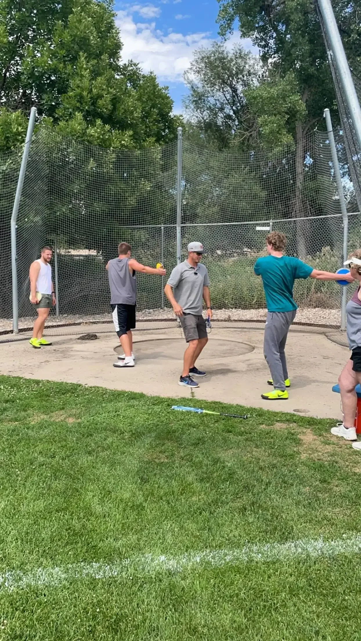 A group of young athletes practicing discus on an outdoor field, with a netted cage in the background and a grassy area in the foreground.