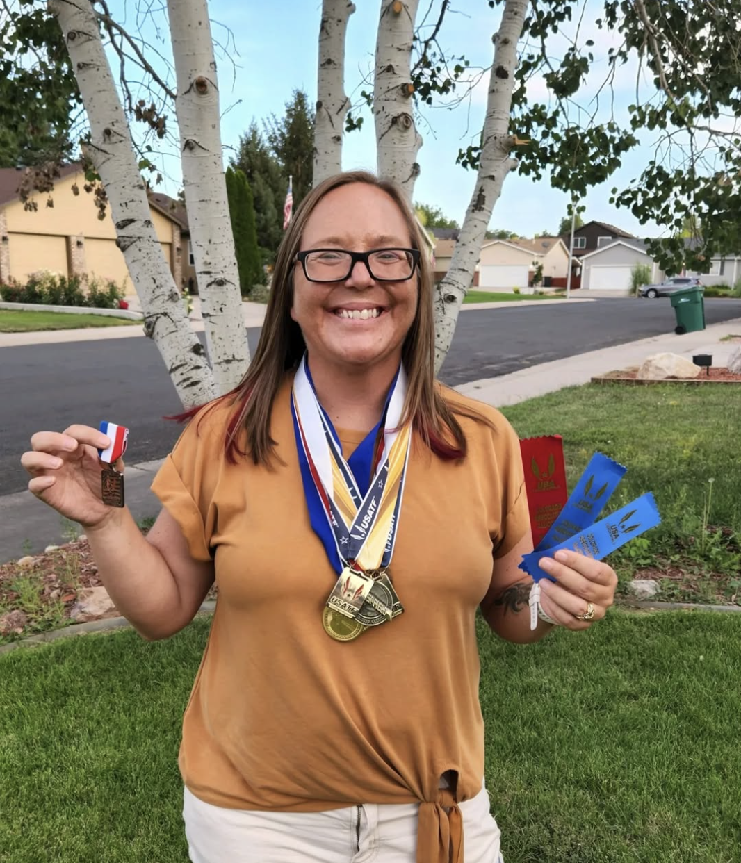A smiling woman with glasses and long brown hair holds medals and ribbons in a suburban yard. She is wearing a tan shirt, white shorts, and has medals around her neck, celebrating an athletic or competitive achievement.