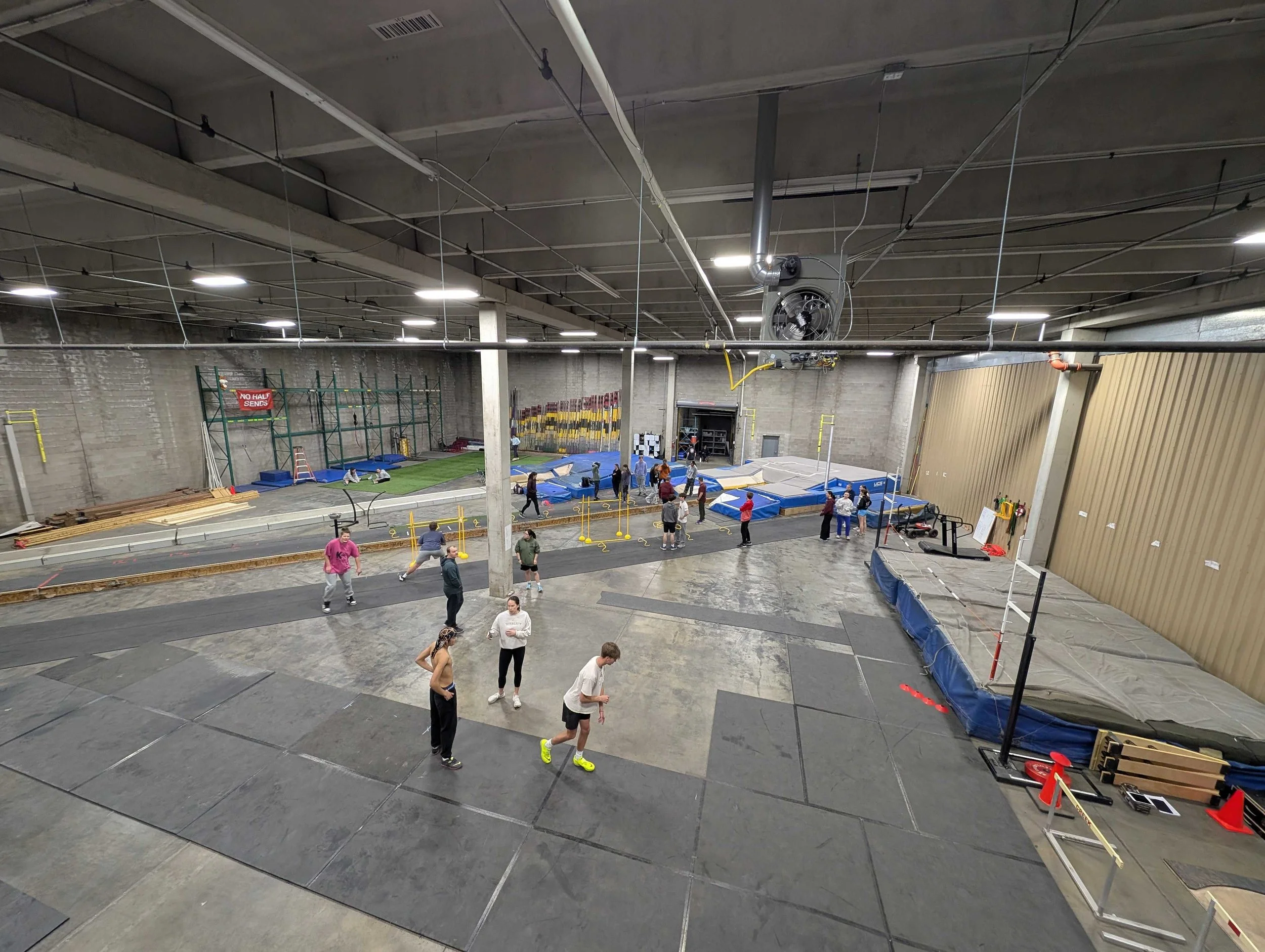 Indoor track and field training facility with multiple zones, people jumping and walking, and fitness equipment, with a high ceiling and industrial design.