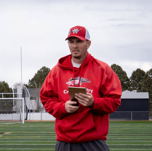A man in a red sports hoodie and cap standing on a track field, holding a tablet.