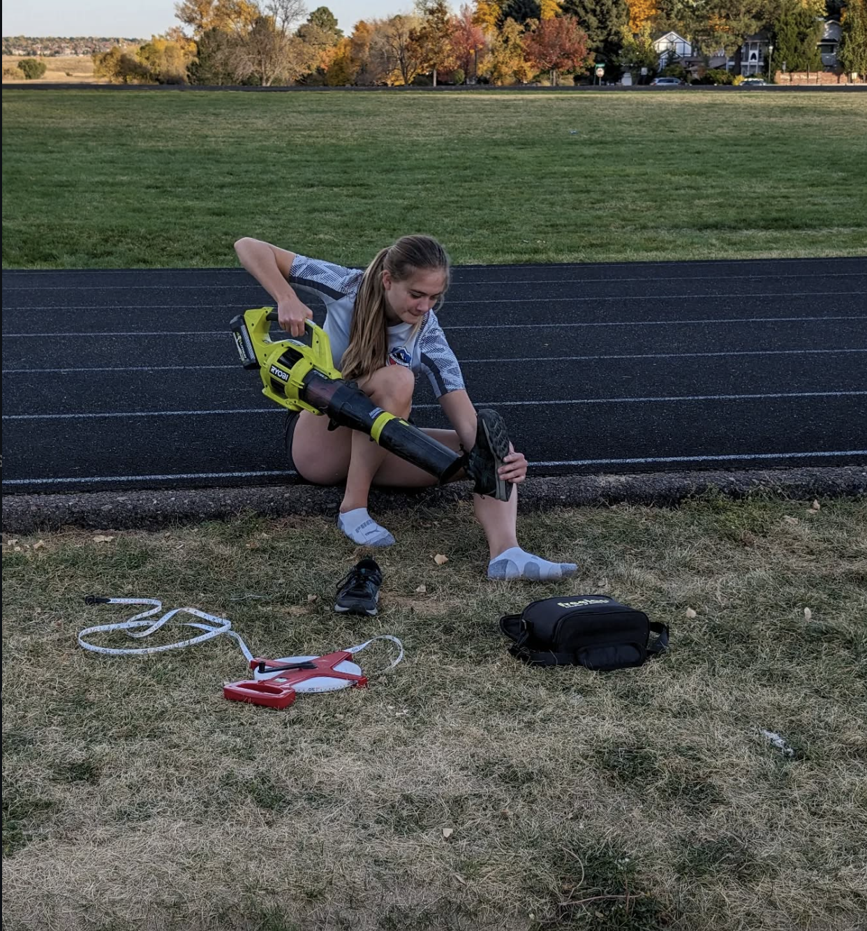 A young girl in athletic clothing using a leaf blower to clear debris from a track at an outdoor sports field, with a black bag and a red and white device nearby, and trees with fall foliage in the background.