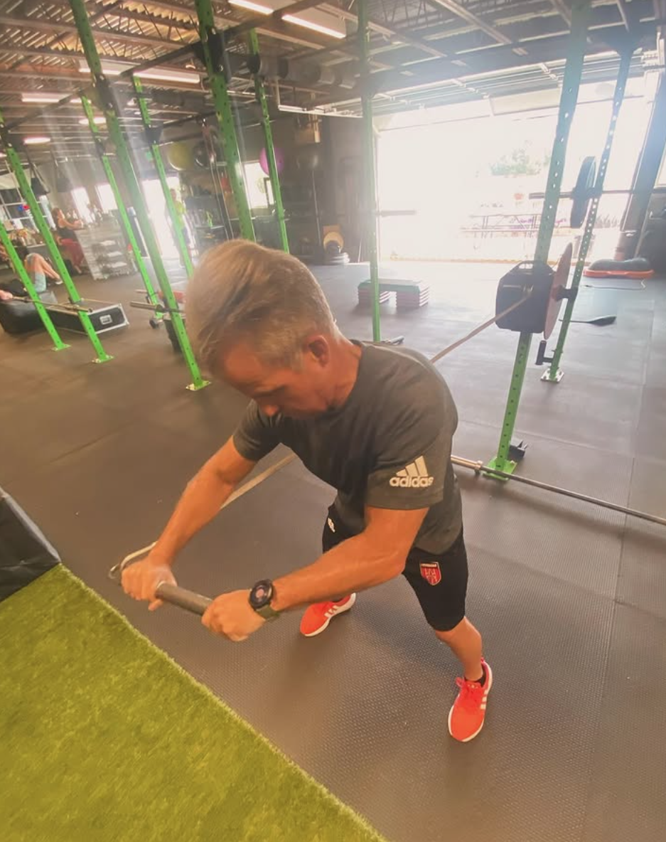 An older man with gray hair working out with a battle rope in a gym. He is leaning forward and pulling the rope with both hands, wearing a black Adidas t-shirt, black shorts, red sneakers, and a watch.