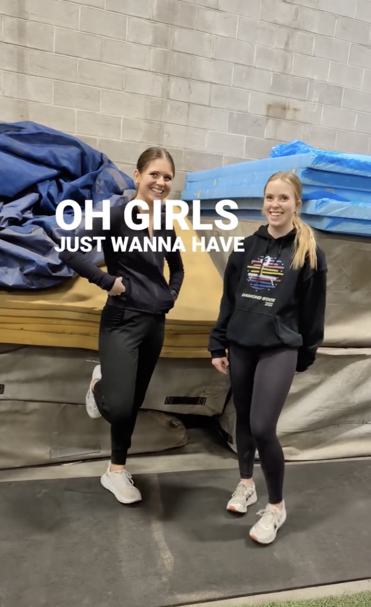 Two women smiling and standing in an indoor sports or gym storage area, surrounded by mats and foam padding, with a quote overlay that says "OH GIRLS JUST WANNA HAVE".