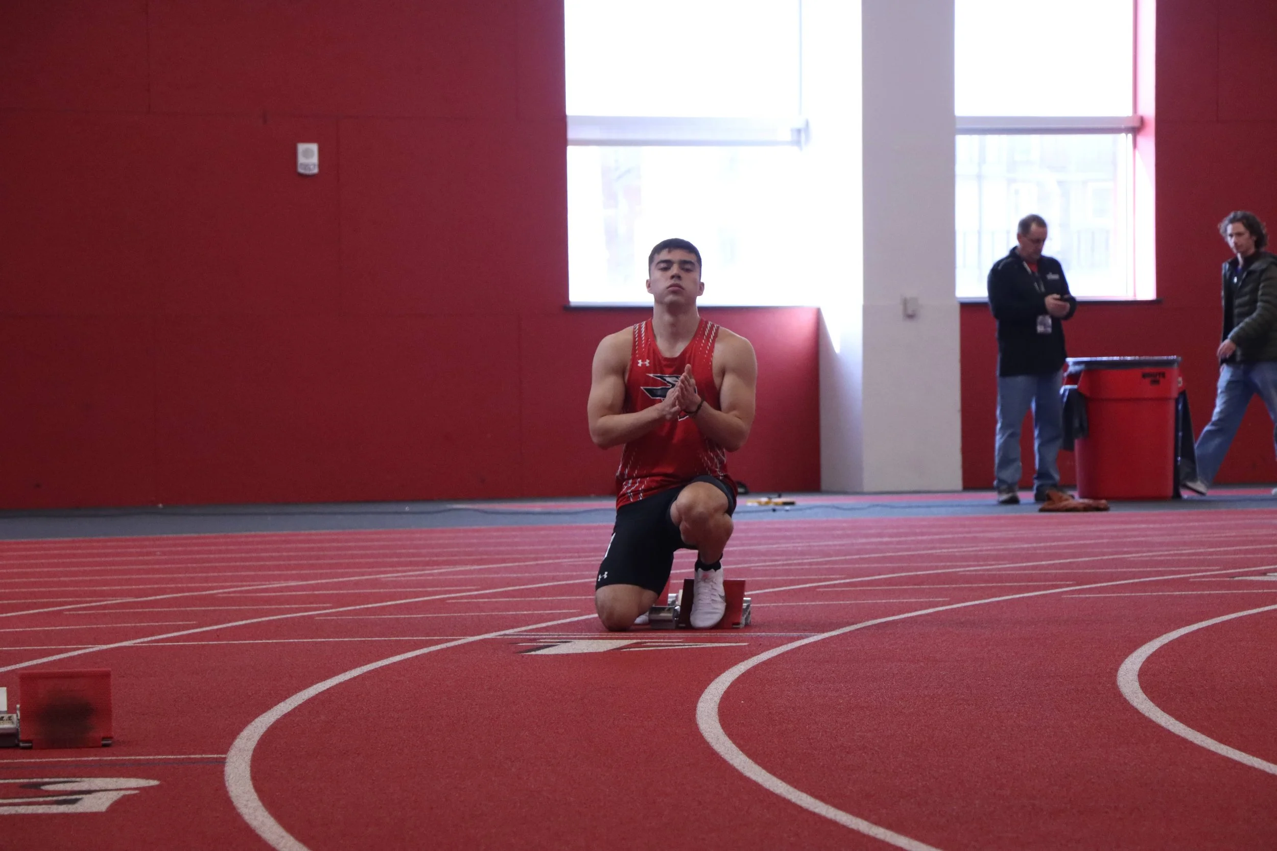 Male athlete kneeling on a running track, preparing for a race, inside a sports facility with red walls and large windows, with two people in the background near a red trash bin.