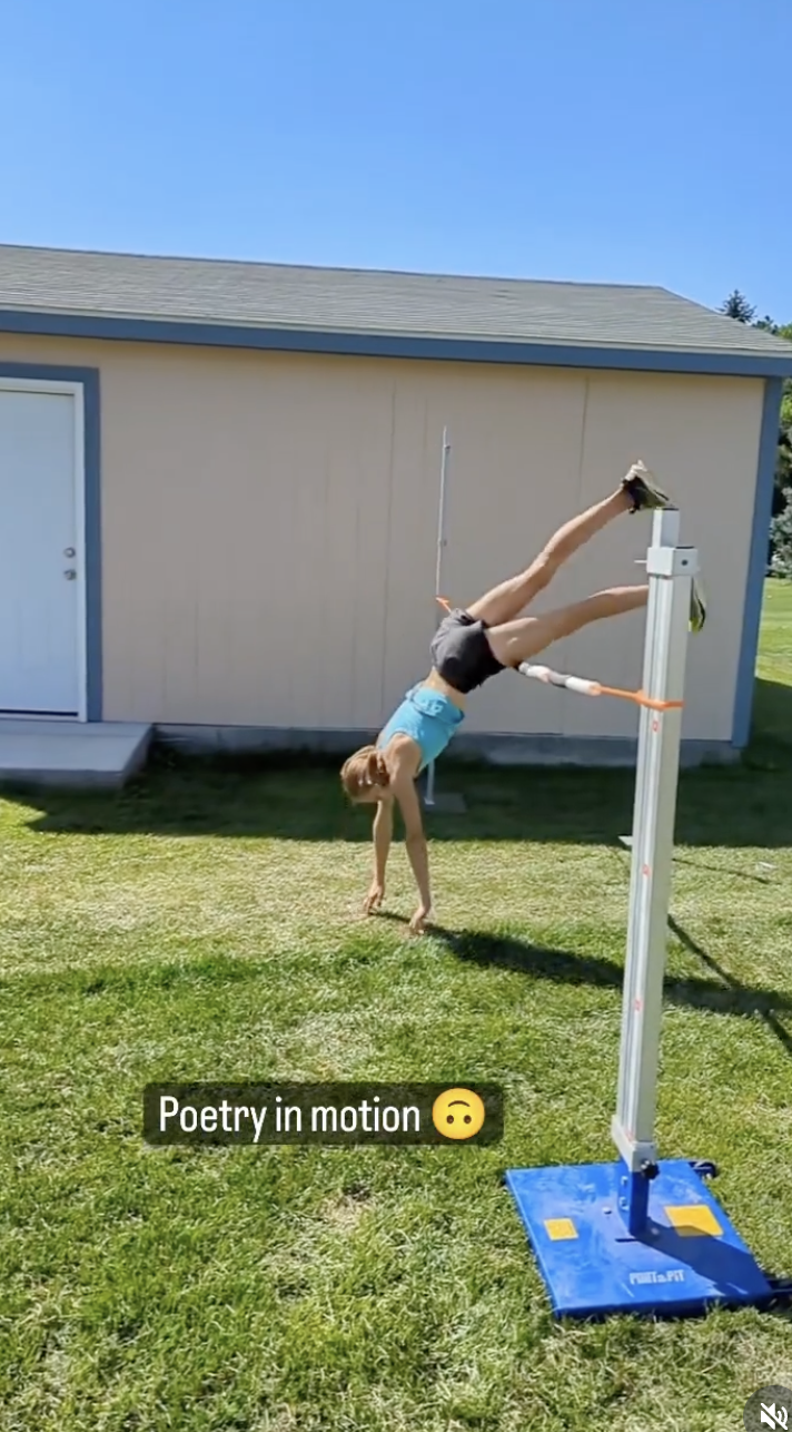 A person performing an acrobatic handstand on a horizontal bar outside, with the body bent at the waist and legs extended upward. The person is wearing a turquoise tank top and black shorts, on a grassy area near a beige shed or small building. The s