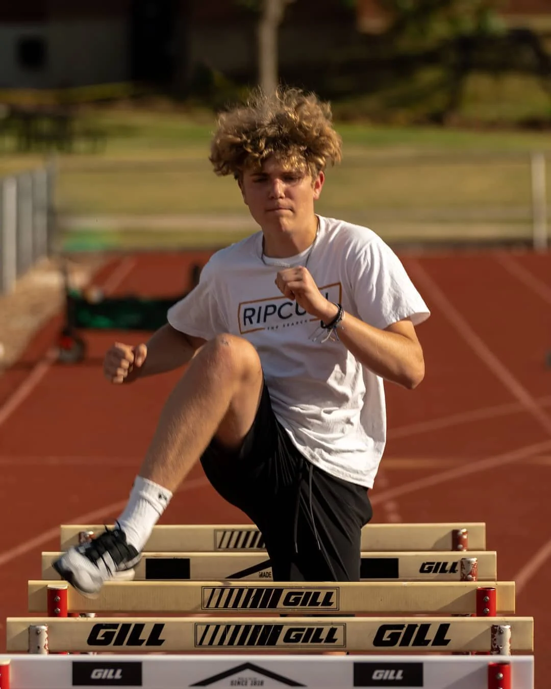 A young man with curly hair is running and jumping over hurdles on an outdoor track.