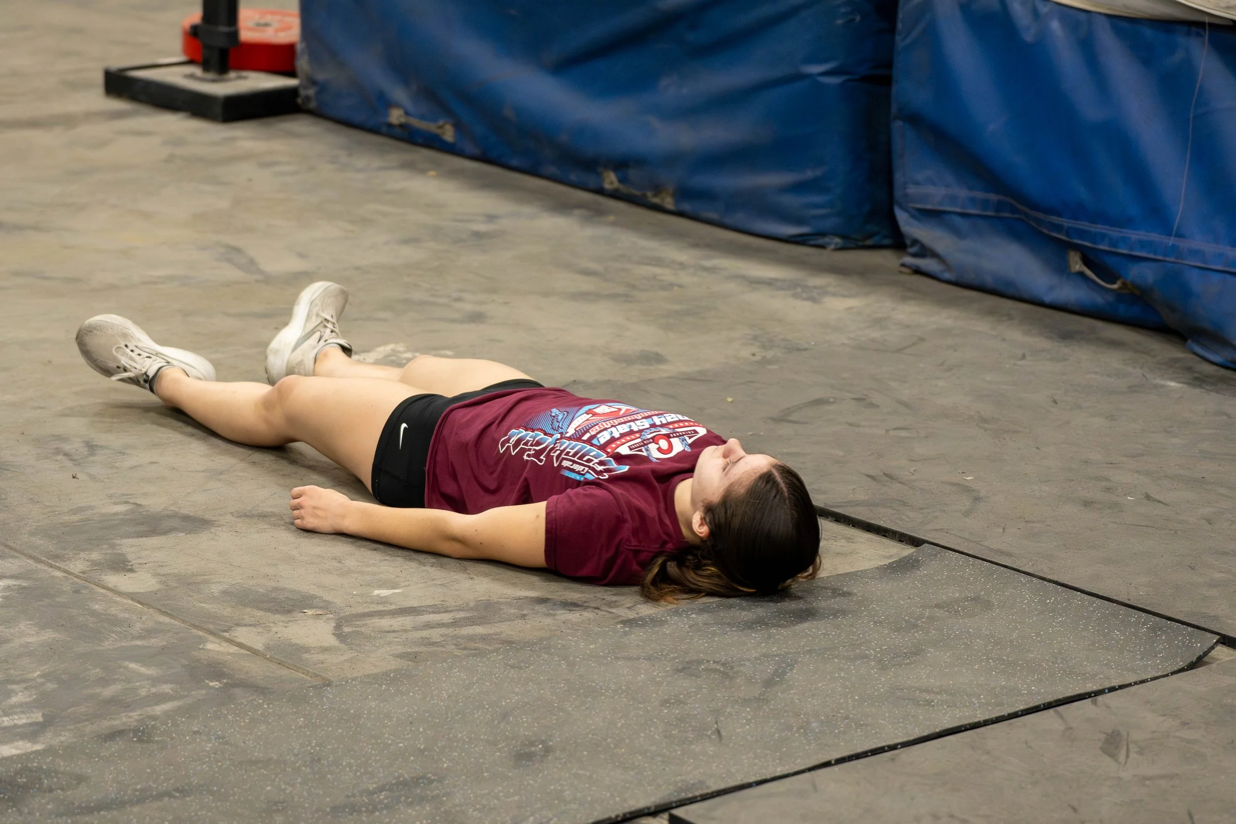 A young woman lying on her back on a gym floor, appearing to be resting or exhausted, wearing athletic clothes and sneakers.