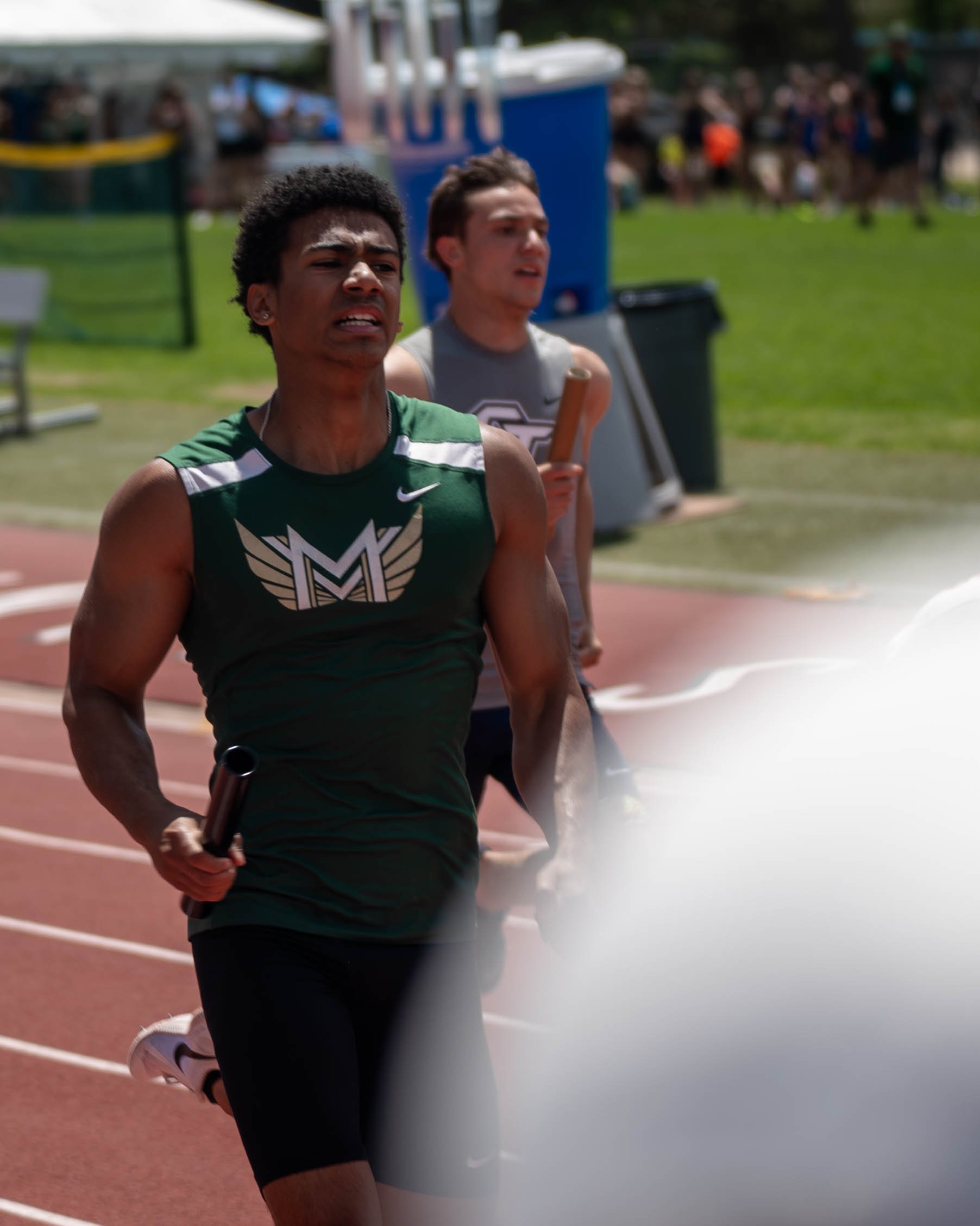 Two male athletes participating in a relay race on a track, with the one in front holding a baton, and another behind holding a relay baton, with spectators in the background.