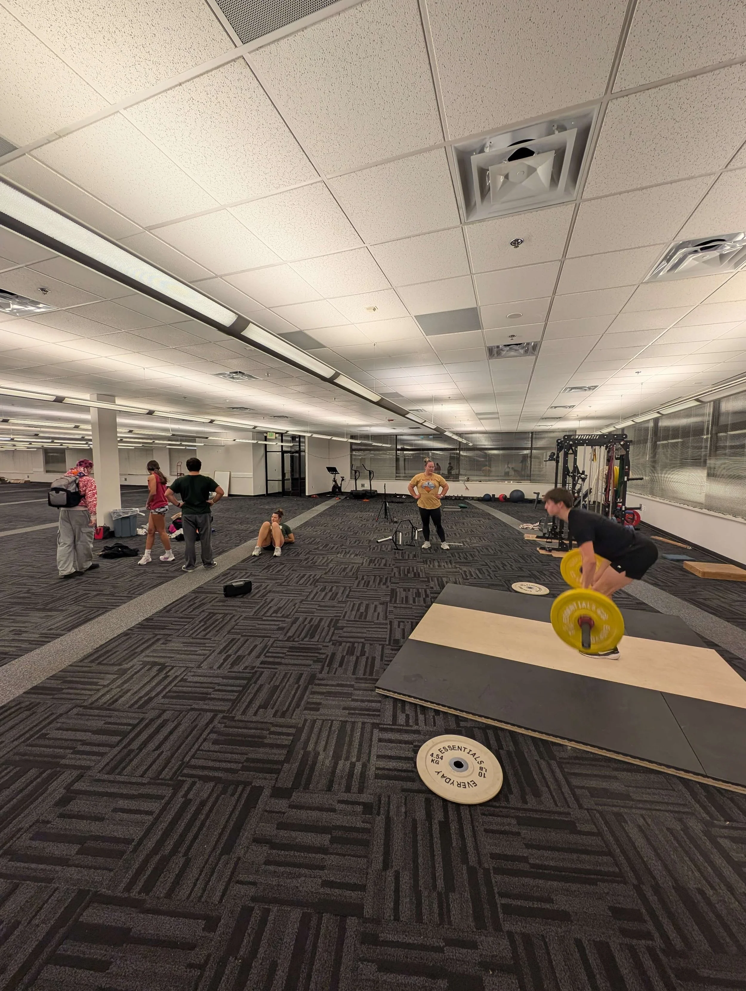 People working out in a spacious gym with black carpeted flooring, including a person lifting weights on a platform, others stretching, and a seated individual, with various fitness equipment in the background.