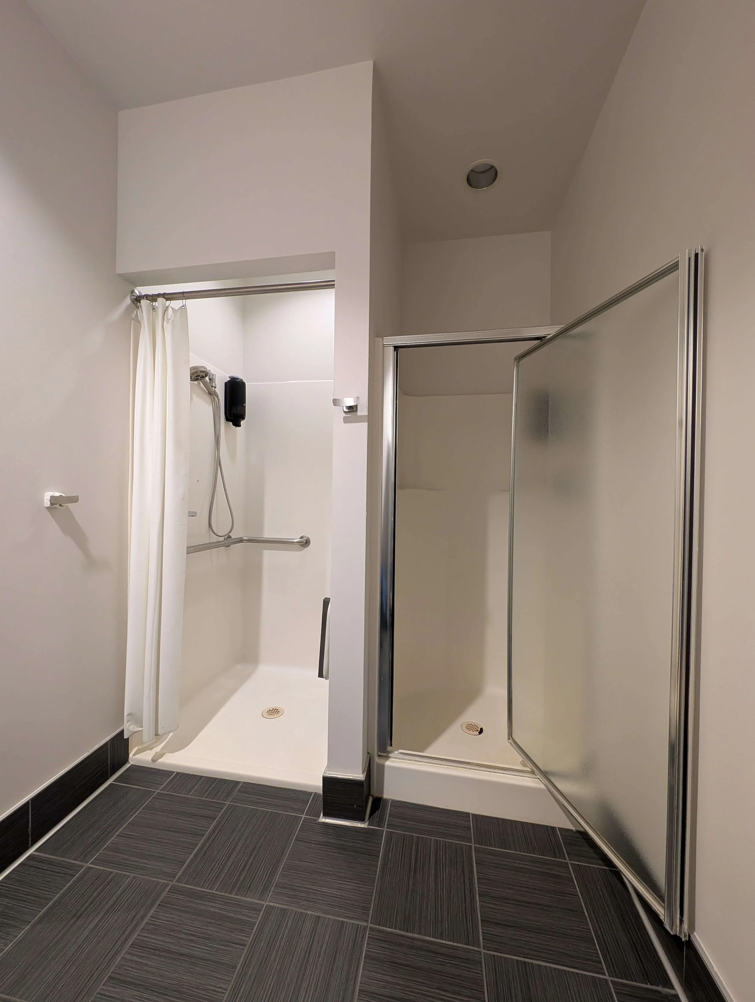 Two shower stalls with sliding glass doors; one with a cream curtain, white walls, and a black shower head, the other with frosted glass. Black floor tiles.