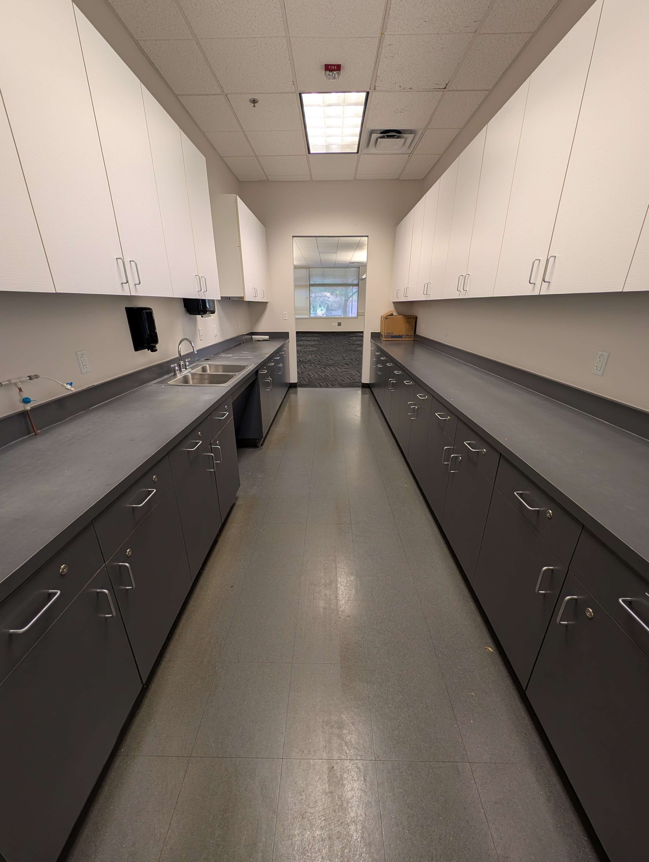 Empty commercial kitchen with black countertops, white upper cabinets, black lower cabinets, stainless steel sink, and a doorway leading to a well-lit room.