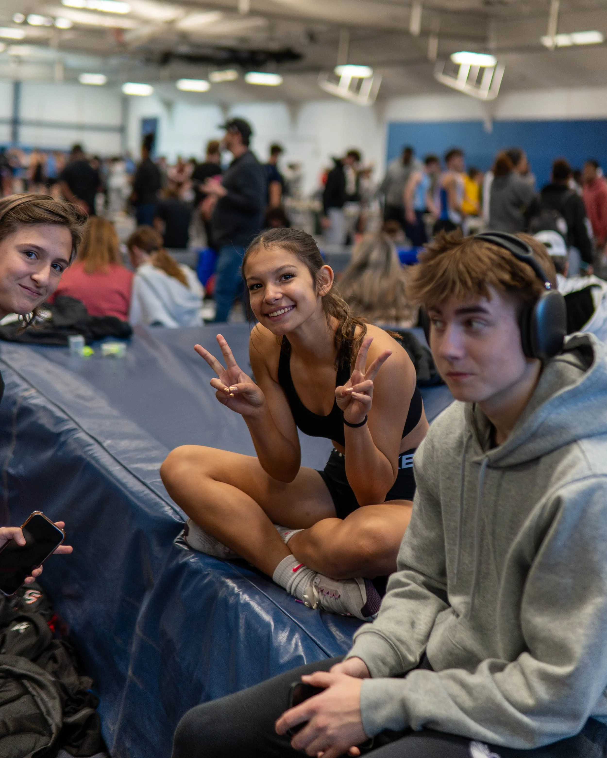 Three young people sitting on blue padding in a crowded indoor gym, with a large group of people in the background. The girl in the middle is smiling and making peace signs with both hands, wearing a black sports outfit and sitting cross-legged. The boy to her right is wearing gray hoodie and headphones, looking to his left. The boy to her left is smiling at the camera, holding a phone.