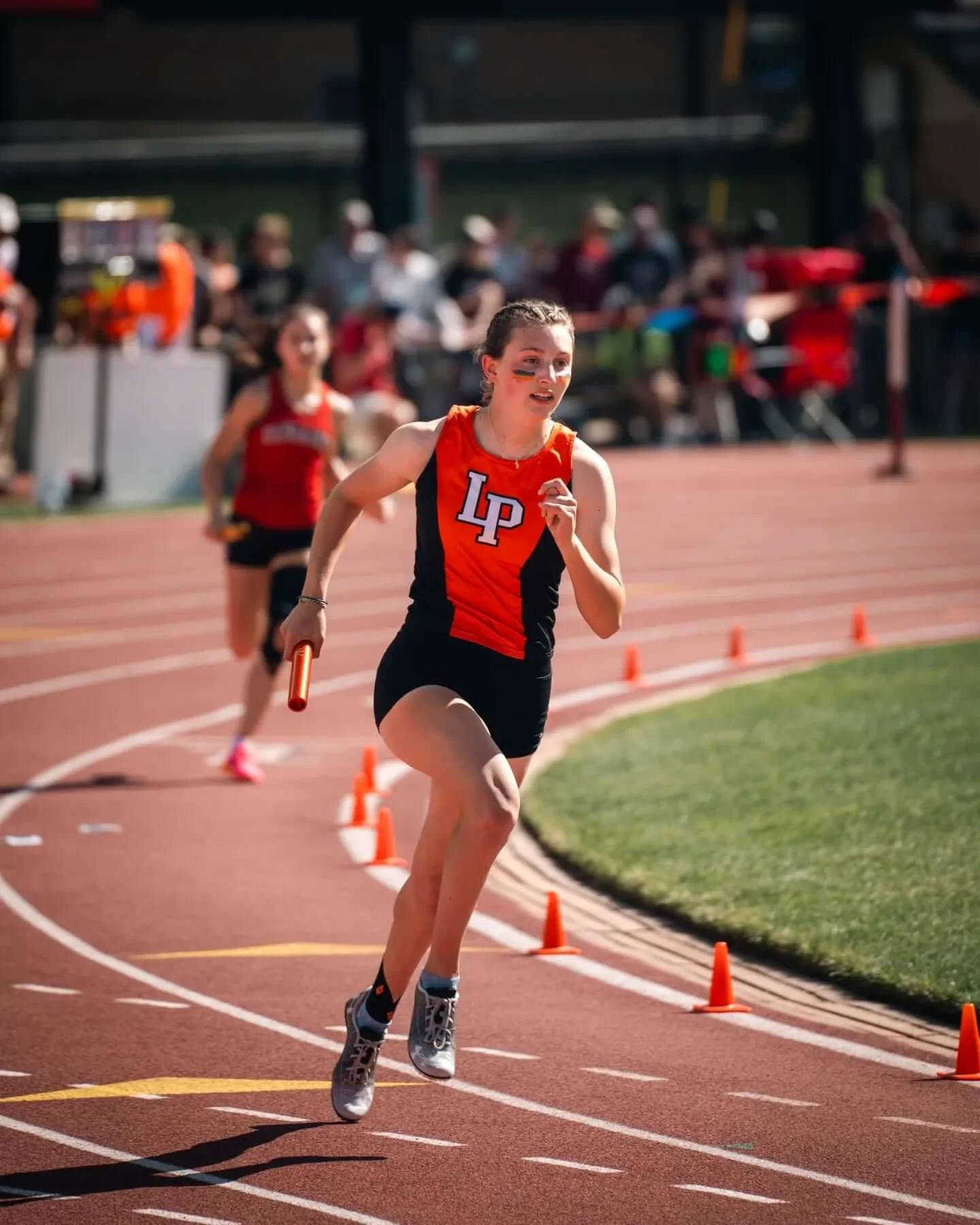 A young female athlete running on a track during a relay race, holding a baton. She is wearing a black and red sports uniform with 'LP' on it, and has the number 2 on her arm. There are orange cones along the track and spectators in the background.