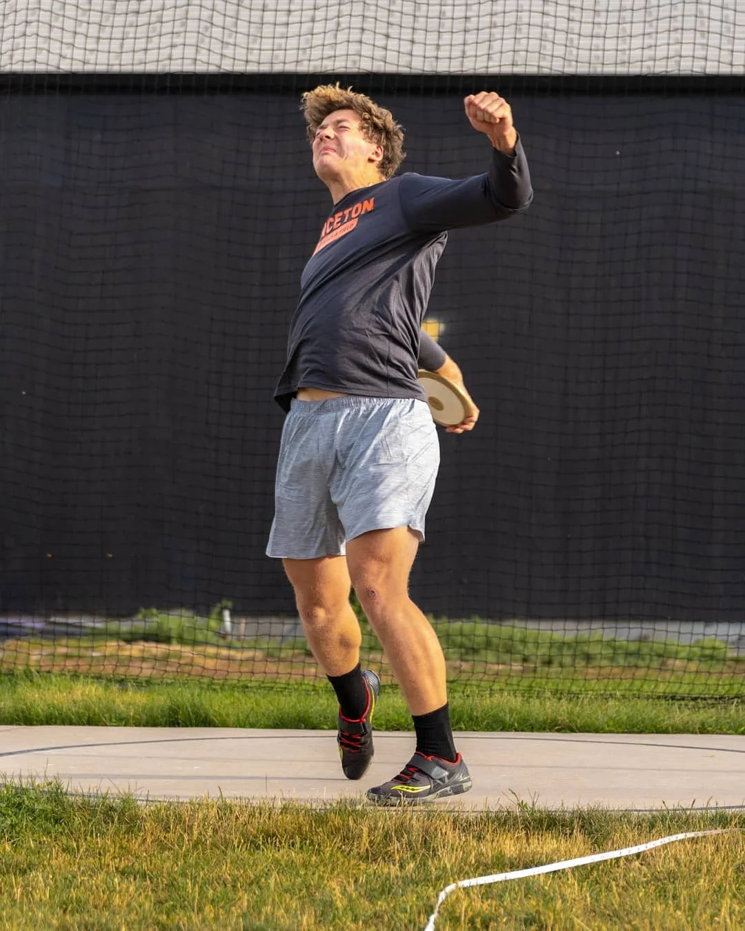 A young man in athletic clothes is celebrating after throwing a discus, standing on a track with a sports field and netting in the background.