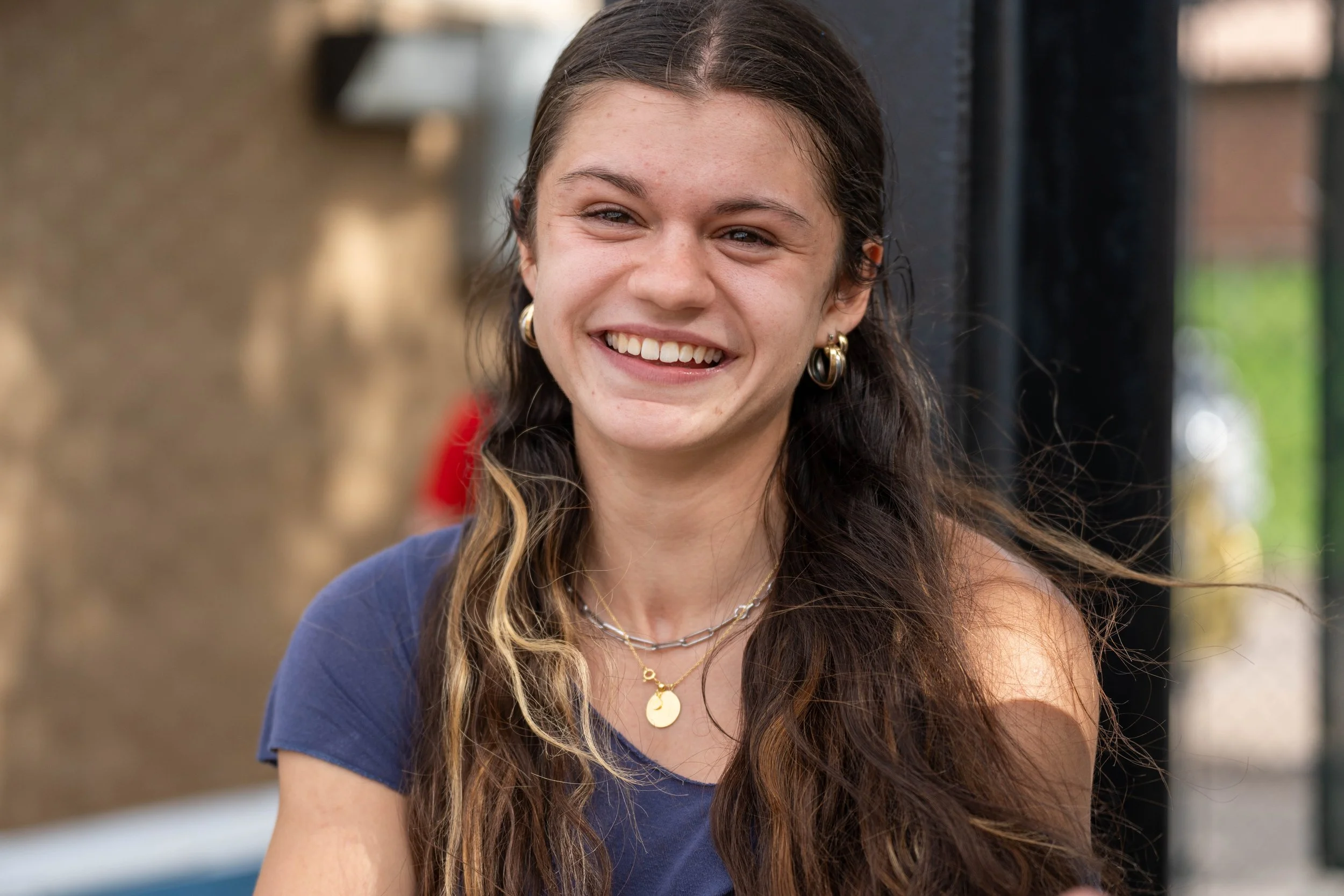 A young woman with long wavy brown hair smiling outdoors, wearing gold hoop earrings, layered necklaces, and a blue top.