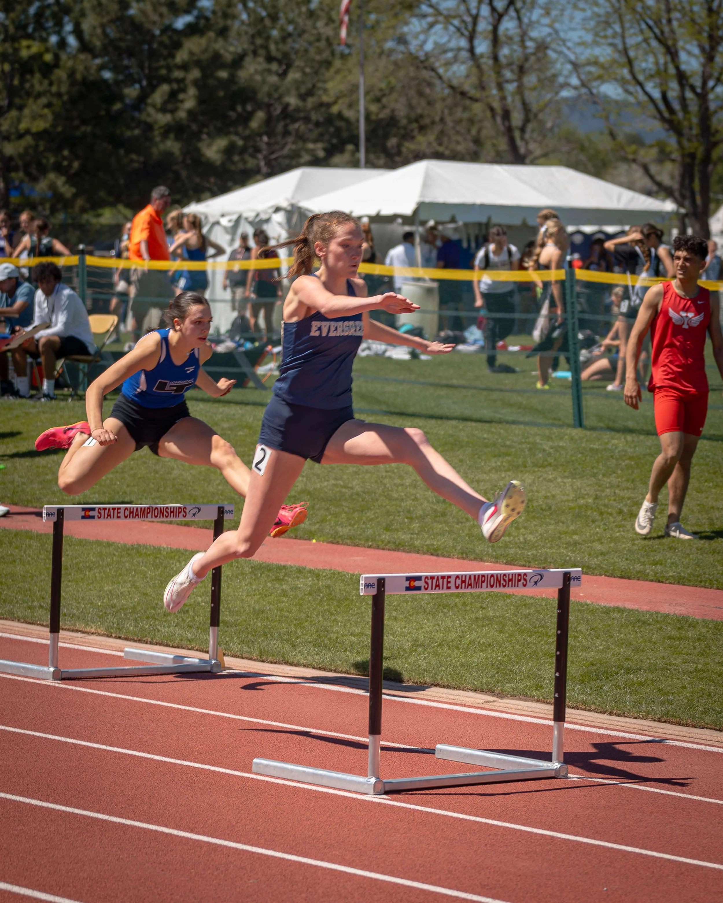 Girls competing in a hurdle race during track and field event at a stadium on a sunny day.