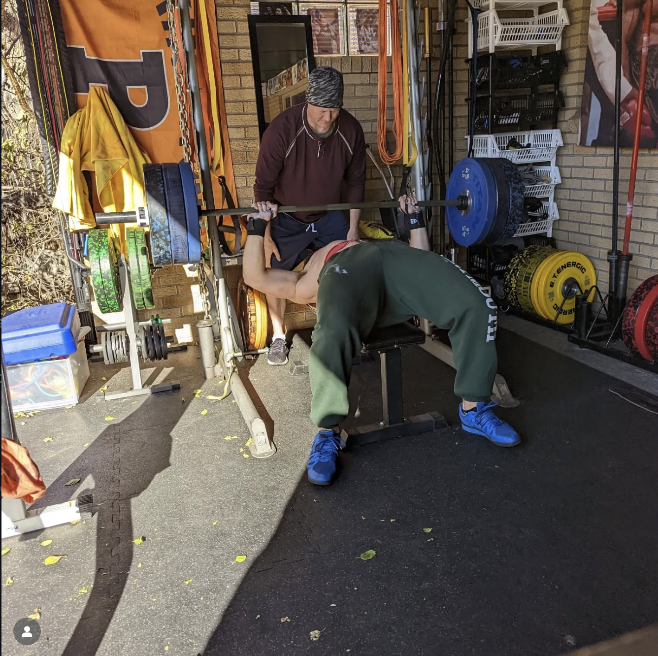 A man spots a woman bench pressing weights in an outdoor gym area, with various gym equipment and weights stored on shelves behind them.