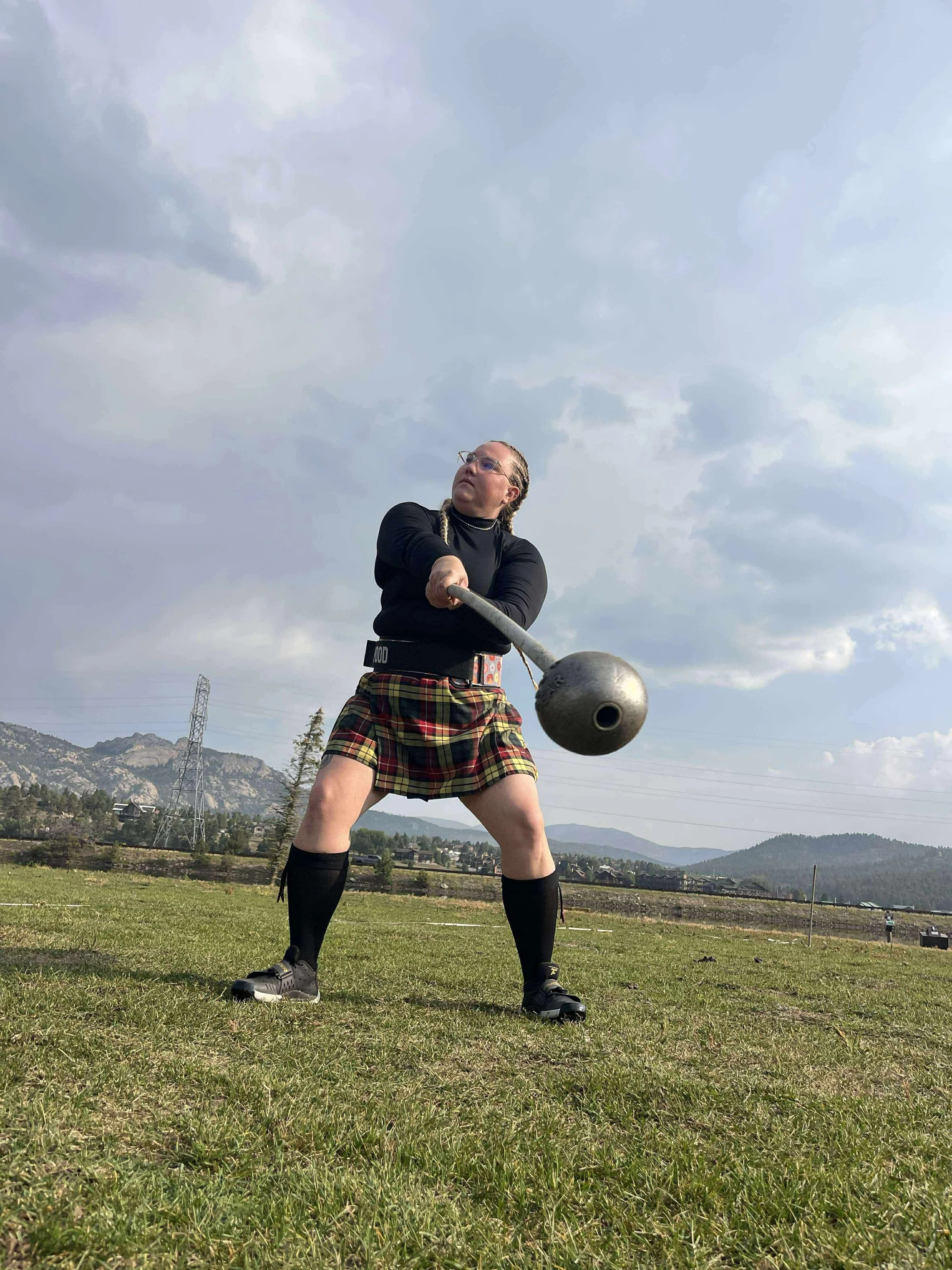 Person wearing a black long-sleeve top, plaid kilt, and black knee-high socks, holding a medieval mace, standing on grass with hills and cloudy sky in the background.