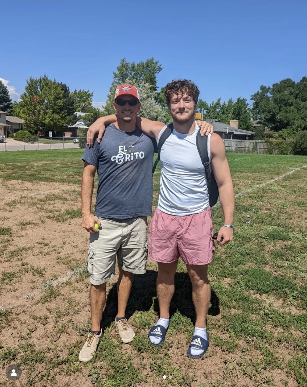 Two young men standing outdoors on a grassy field with arms around each other's shoulders, smiling at the camera, in a sunny day with clear blue sky.