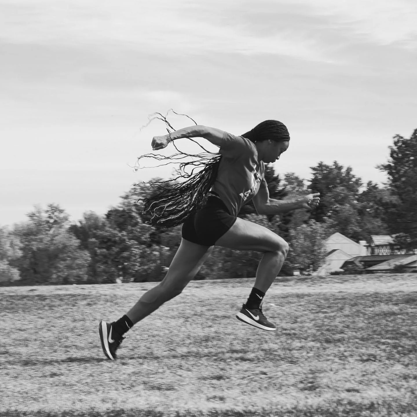 Black and white photo of a woman with long braids running outdoors on a field with trees and sky in the background.