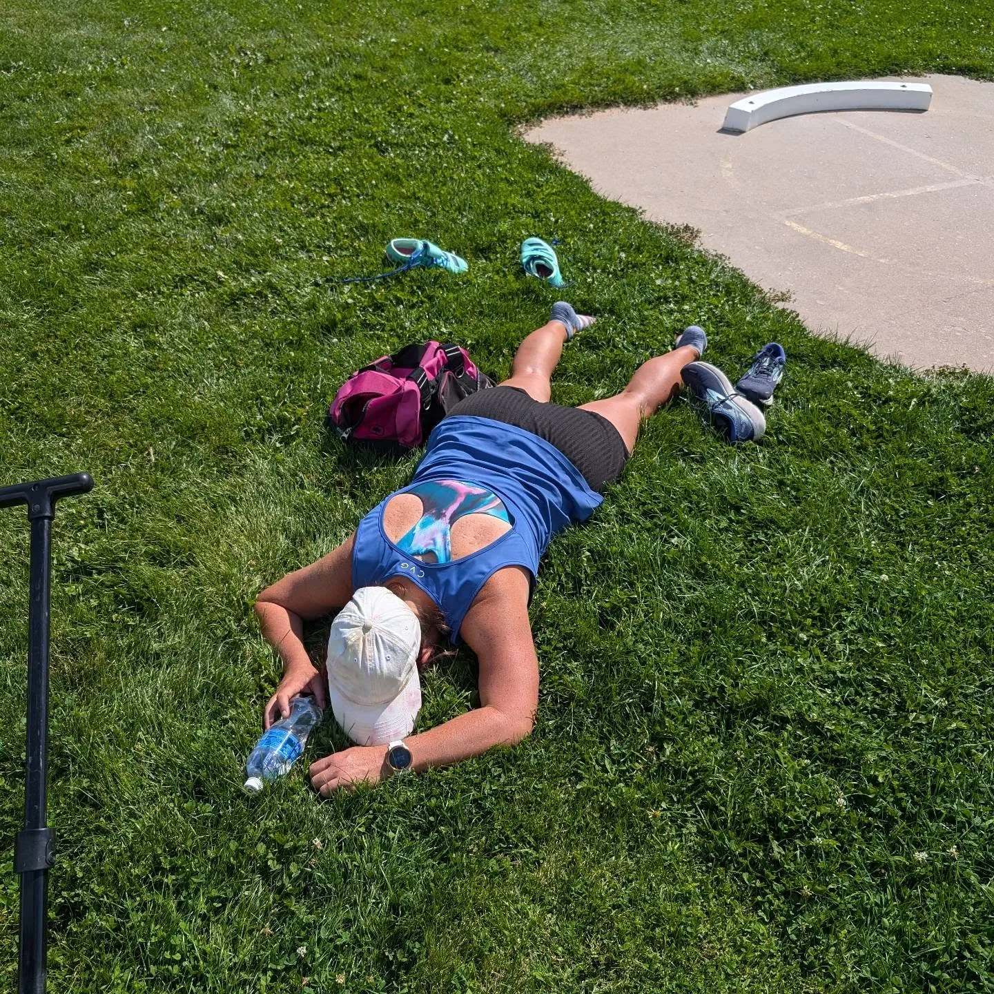 A woman lying on grass after a run, wearing athletic gear, with water bottles, shoes, and a backpack nearby, near a parking lot.