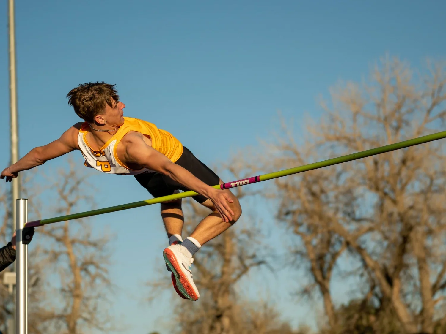 A male pole vaulter in a yellow and white uniform attempting to clear a horizontal bar during a track and field event outdoors, with leafless trees in the background.