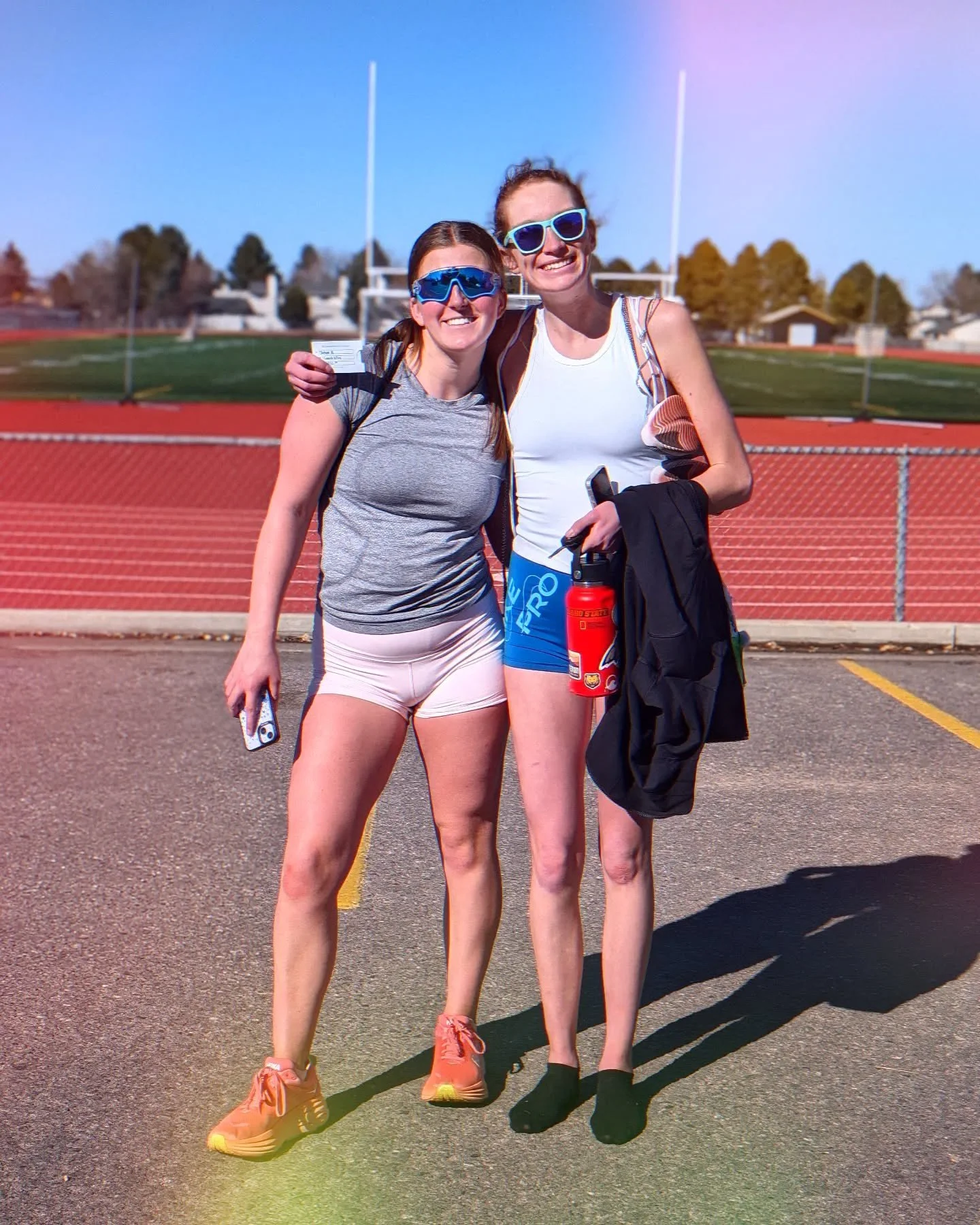 Two women standing side by side on an outdoor track, smiling, wearing athletic clothes and sunglasses, with a red water bottle and a phone.