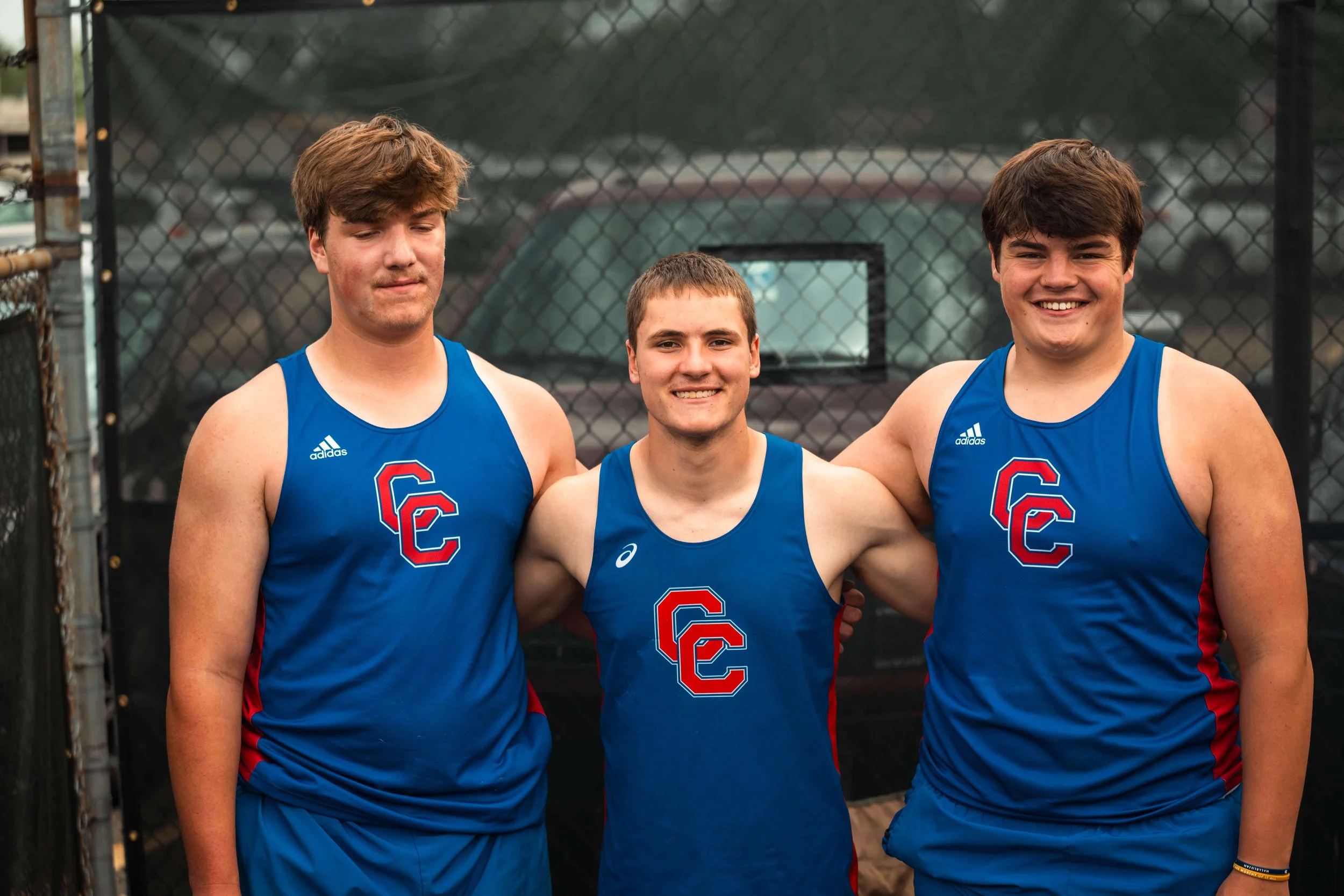 Three young male athletes in blue track uniforms standing together on a track field, with a chain-link fence and cars in the background, smiling.