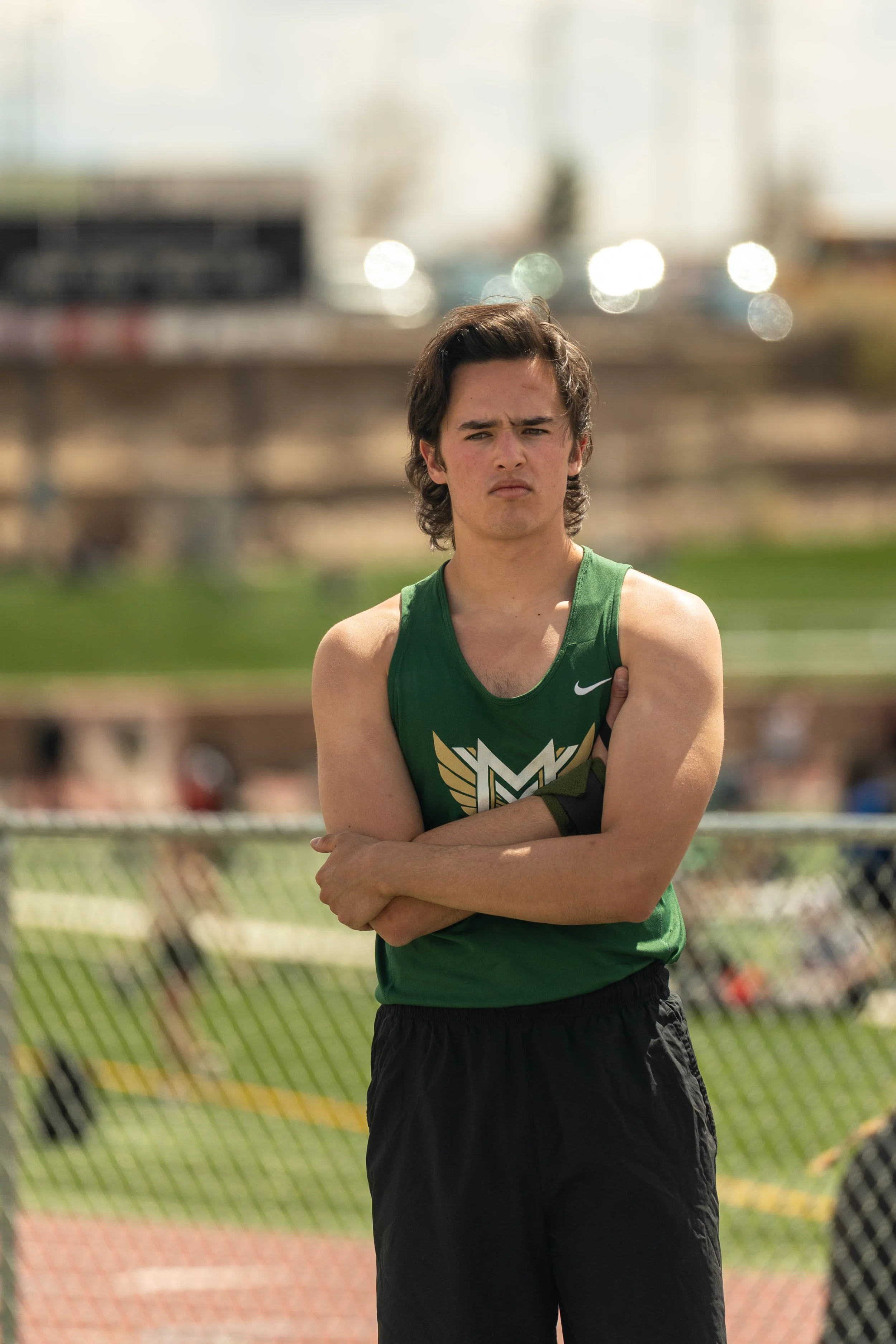 A young male athlete in a green sleeveless sports jersey standing outdoors with crossed arms and a serious expression.