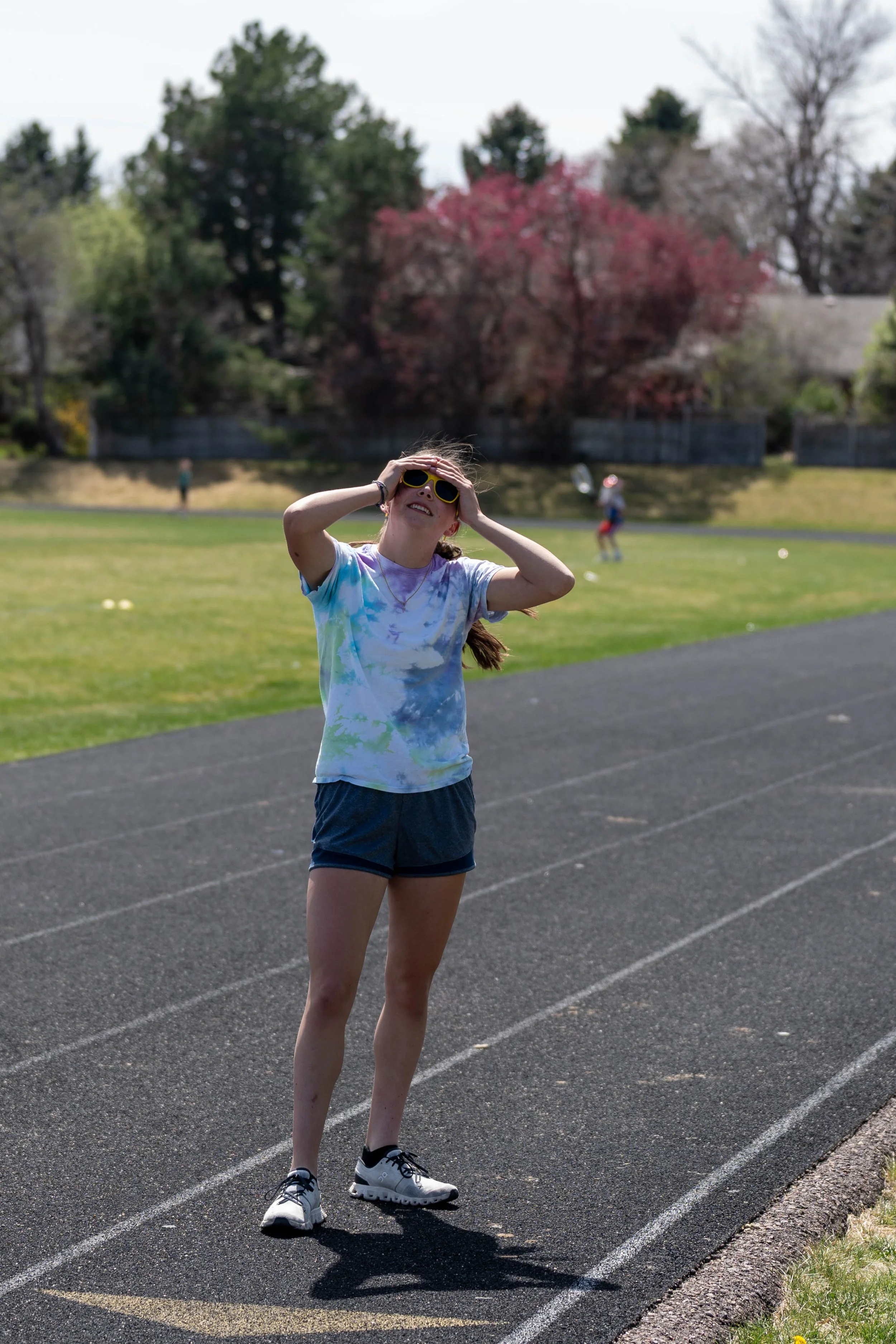 A girl in sunglasses and tie-dye shirt standing on a track, holding her head, with a grassy field and trees in the background, and children playing in the distance.