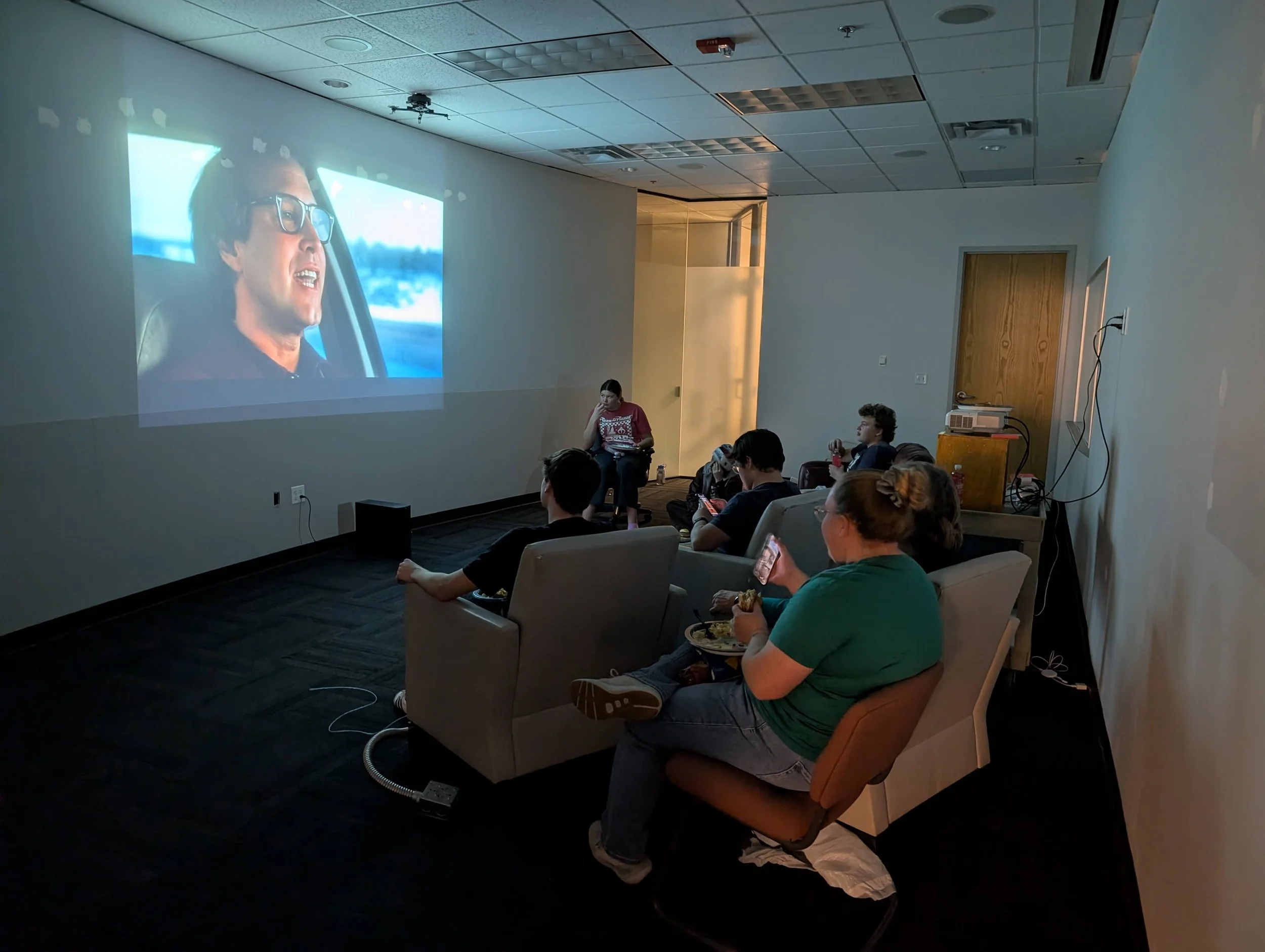 People watching a movie in a small dark room with a large screen showing a man with glasses driving a car.