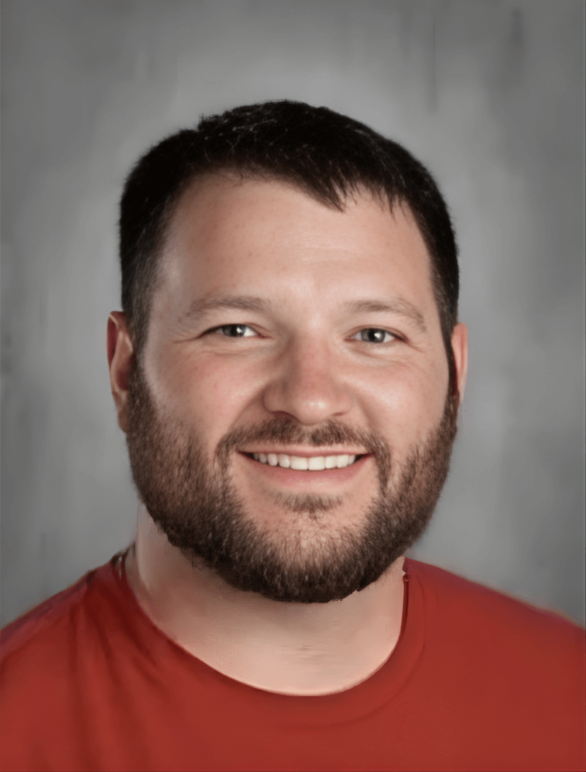 A smiling man with short dark hair, a beard, and wearing a red shirt, posed against a gray background.